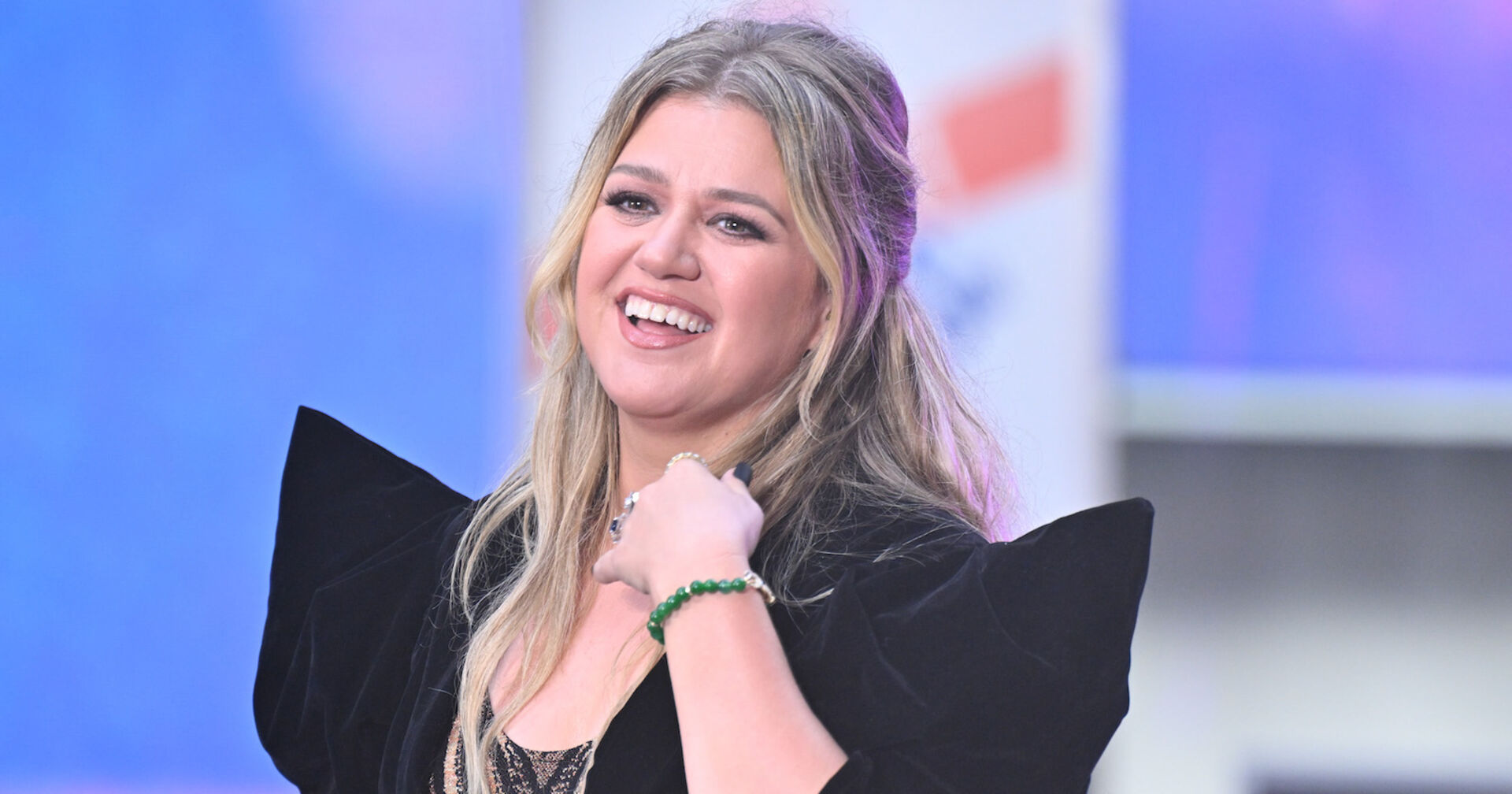 Kelly Clarkson trägt ein schwarzes Kleid und halb geschlossene blonde Haare. Sie lächelt in die Ferne.