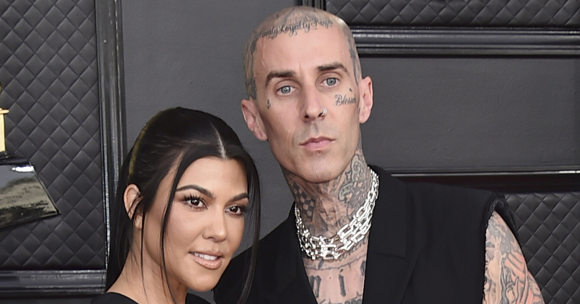 Kourtney Kardashian hat schwarze zugebundene Haare und steht neben Travis Barker, er trägt ein schwarzes Glied, eine silberne Kette und hat unzählige Tätowierungen.