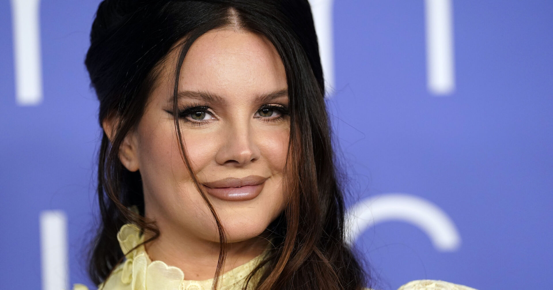 Lana Del Rey trägt hochgesteckte braune Haare und ein starkes Augen Make-Up. Sie steht vor einer blauen Wand.