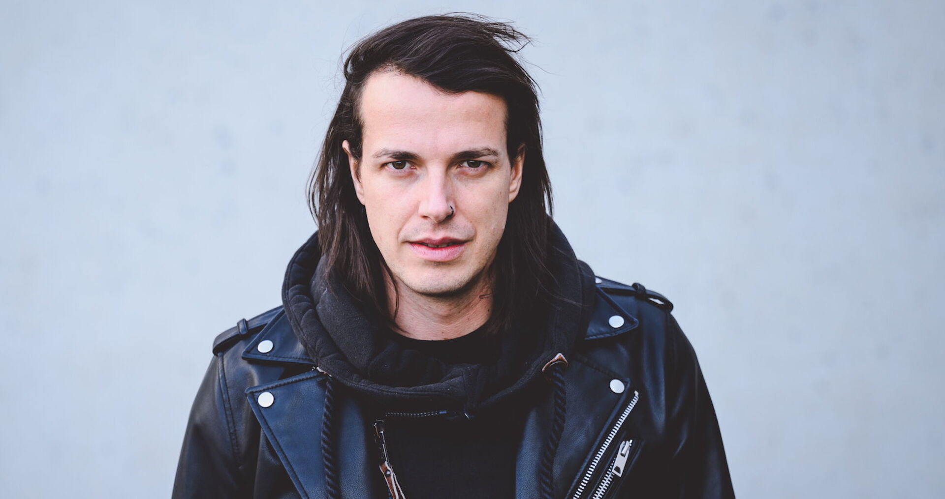 Marco Pogo trägt einen schwarzen Pullover und eine schwarze Lederjacke. Er schaut frontal in die Kamera und steht vor einer grauen Wand.