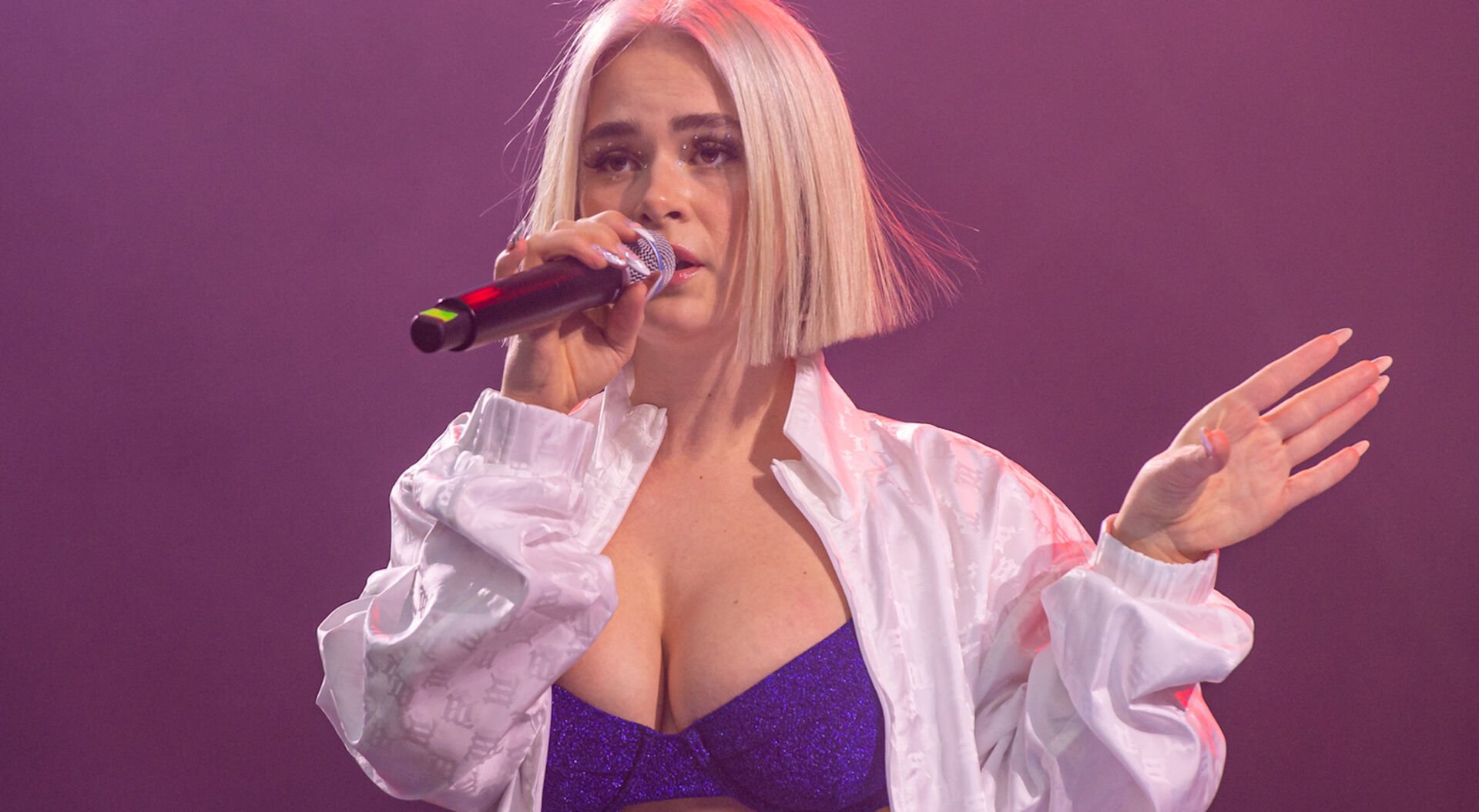 Mathea trägt ein glitzernes Bikinitop und ein weißes Hemd. Sie hat einen blonden Longbob und singt ein Lied.