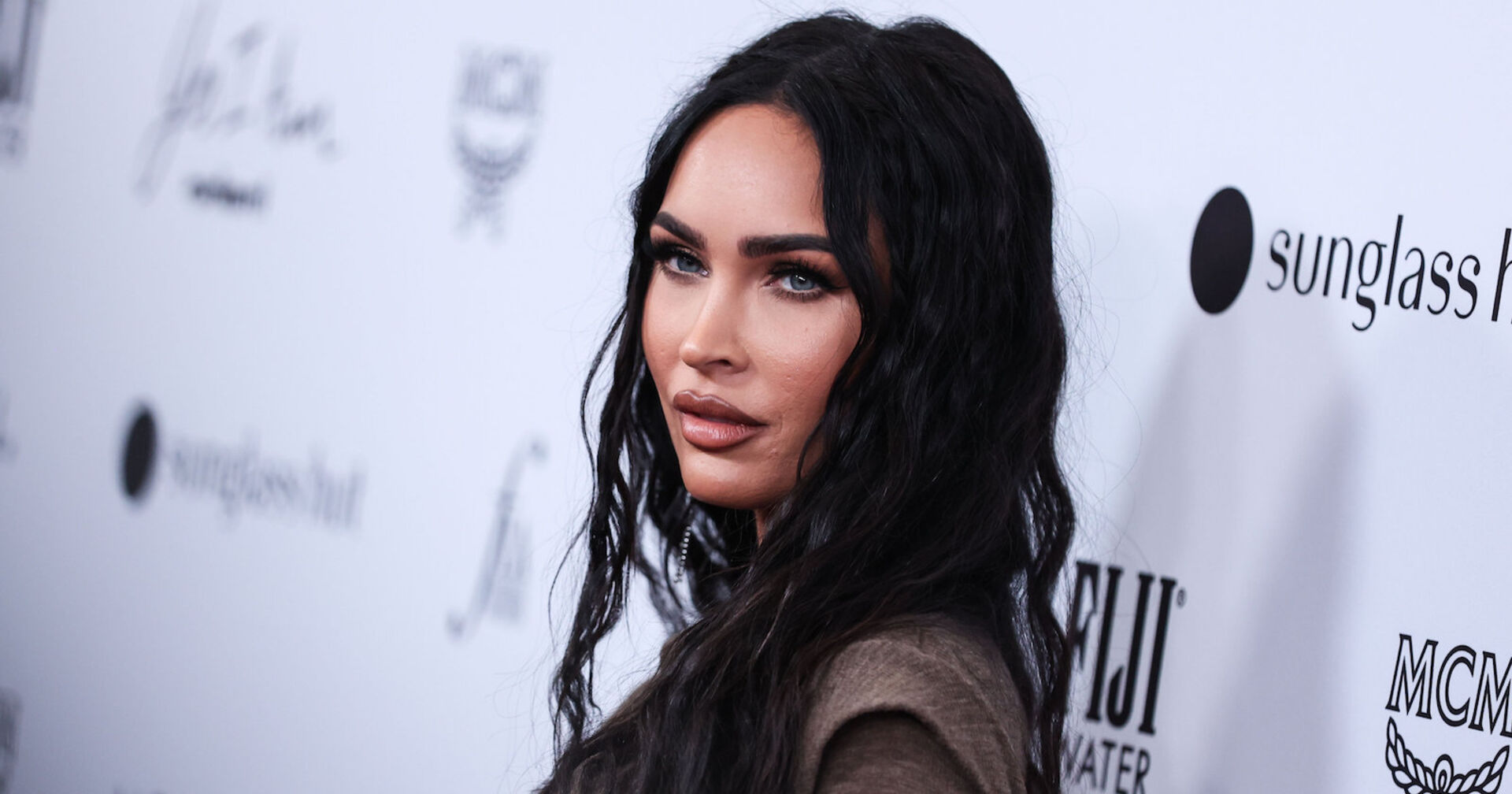 Megan Fox trägt offene schwarze, gewellte Haare und steht vor einer weißen Fotowand.