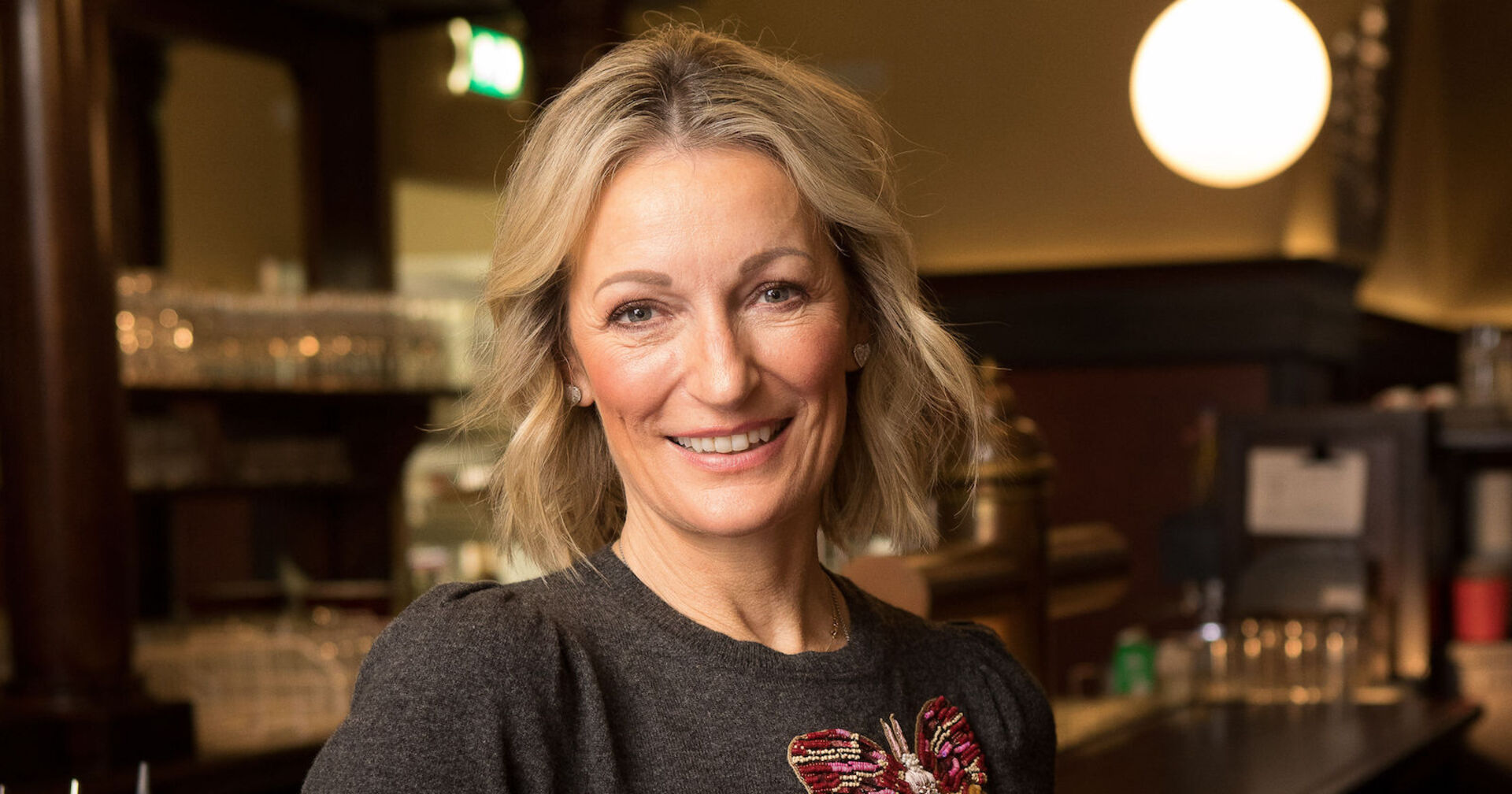 Monika Gruber trägt ein grauen T-Shirt und blonde gewellte Haare in Schulterlange.