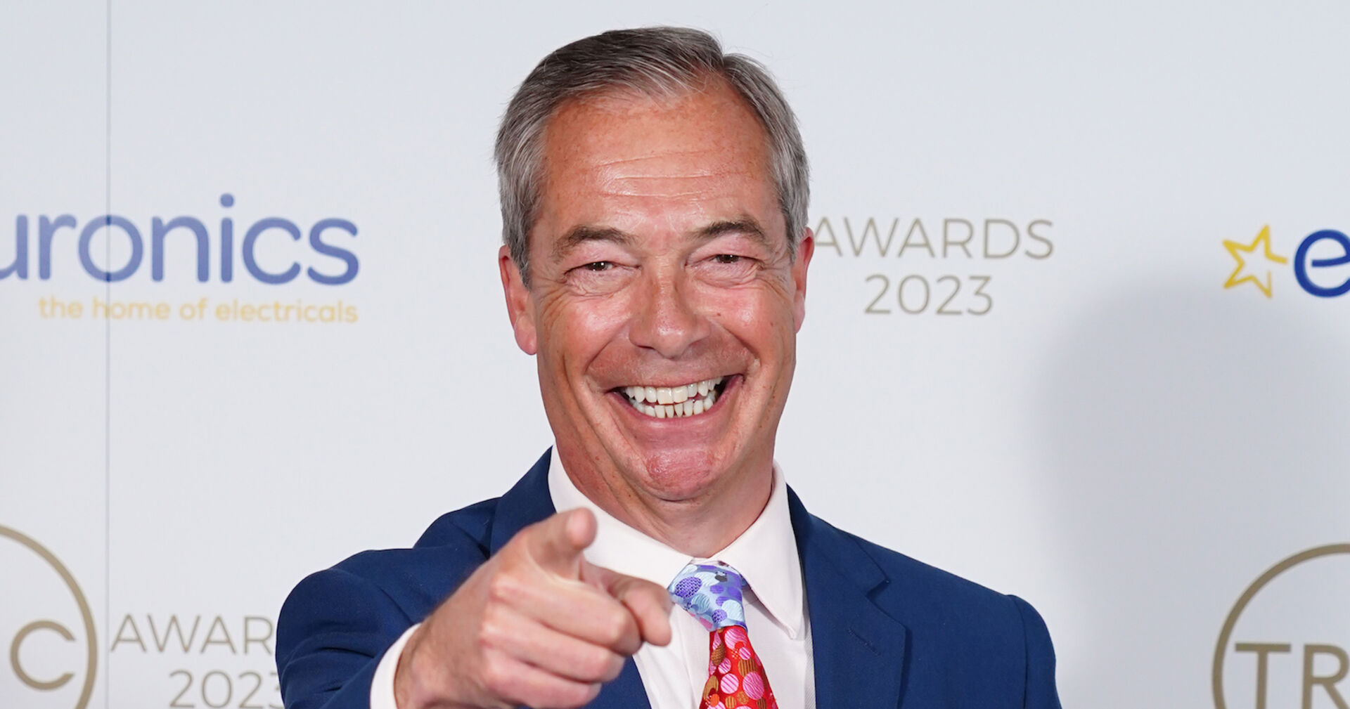 Nigel Farage trägt einen blauen Anzug und eine rote Krawatte. Er grinst in die Kamera und zeigt mit dem Finger auf die Zuschauer