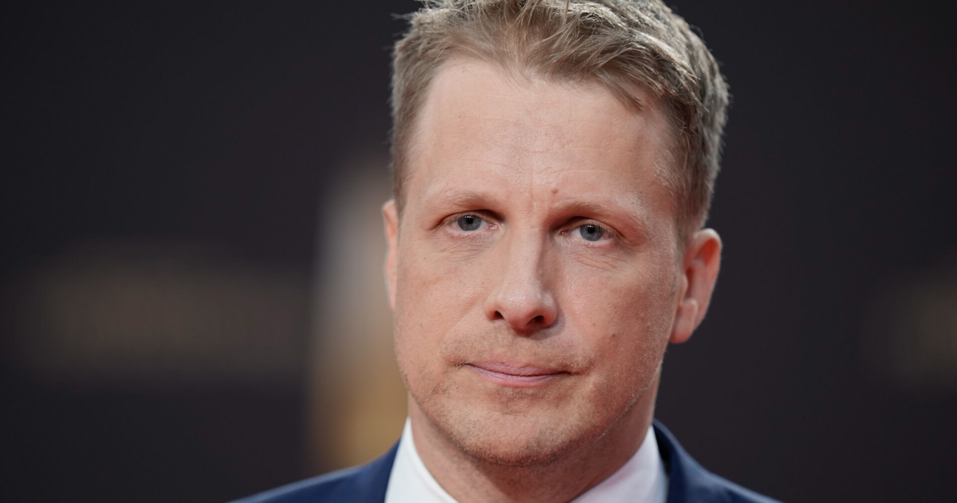 Oliver Pocher trägt einen dunklen Anzug. Er hat kurze blonde Haare und einen traurigen Blick.