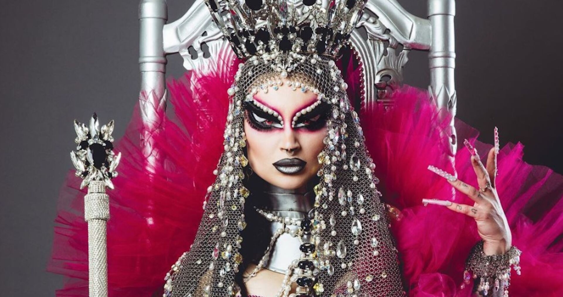 Eine Drag-Queen mit impsanter silvber Krone und pinken Tüll. Sie sitzt auf einem Thron und hält ein Zepter.