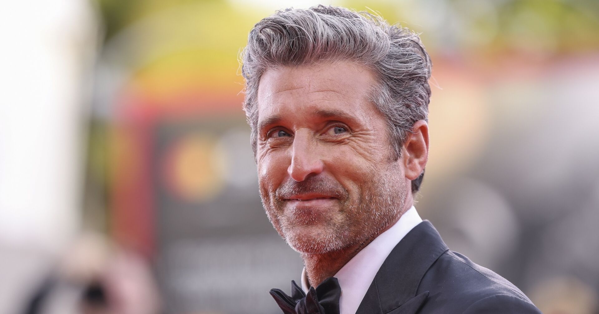 Patrick Dempsey hat graue kurze Haare, einen schwarzen Anzug und ein weißes Hemd. Er blickt von der Seite in die Kamera.