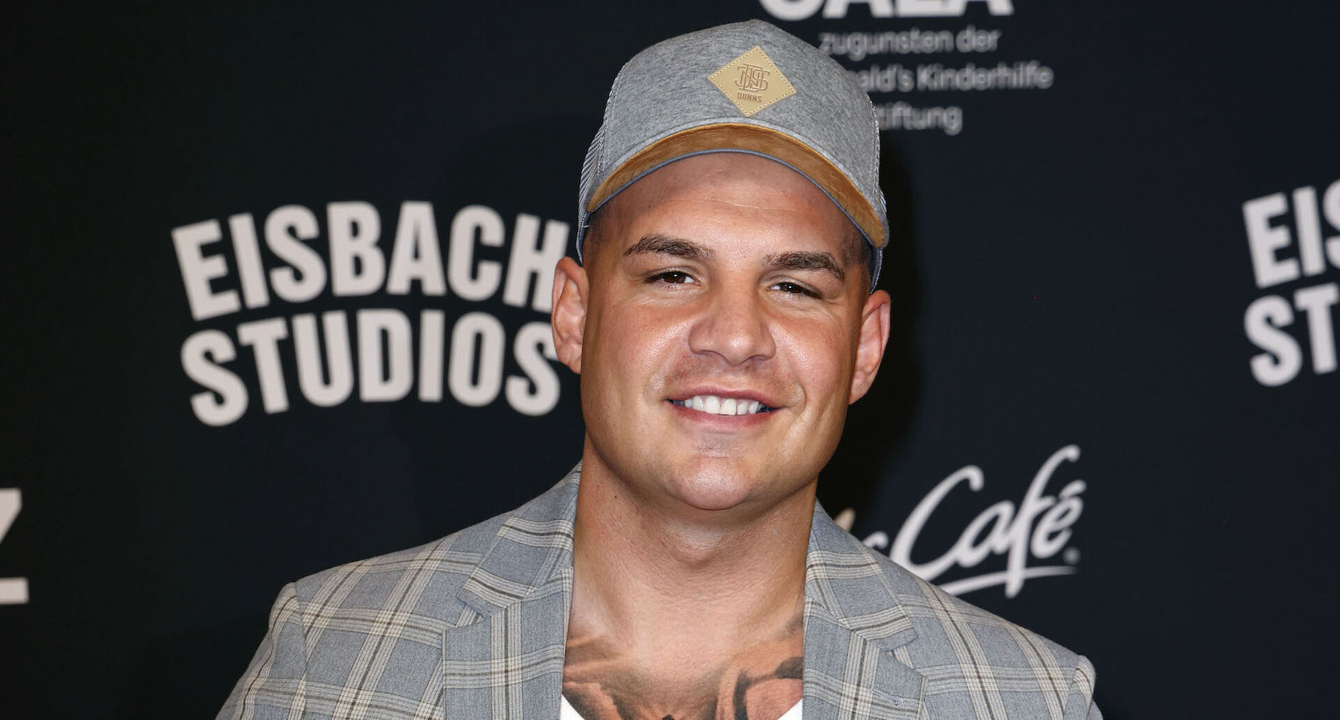 Pietro Lombardi trägt einen karrierten Anzug und eine graue Kappe. Er steht vor einer Fotowand.