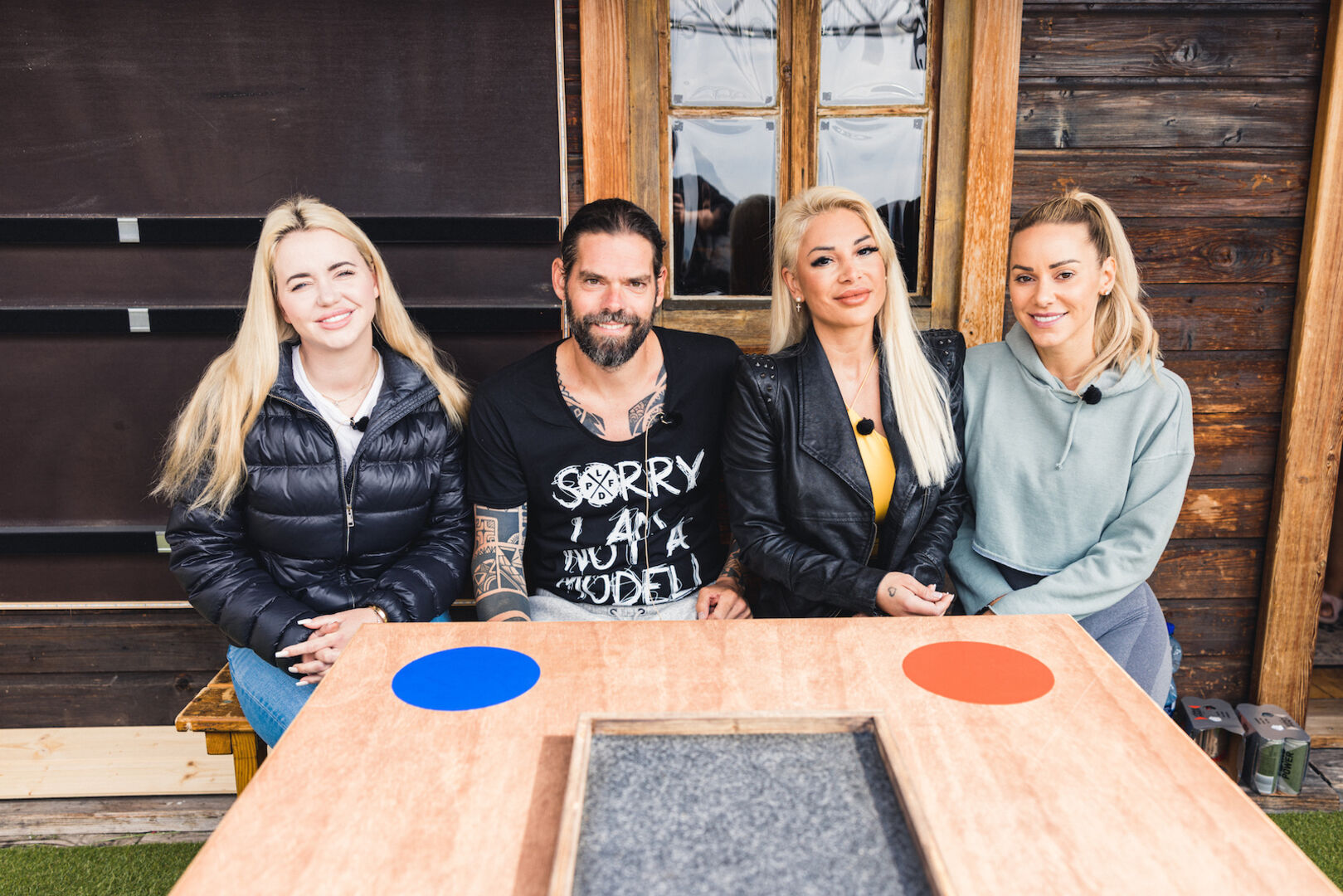Cathy Lugner, Daniel, Sanja und Vanessa sitzen auf einer Holzbank. Vor ihnen ein Spielbrett für die nächste Entscheidung.