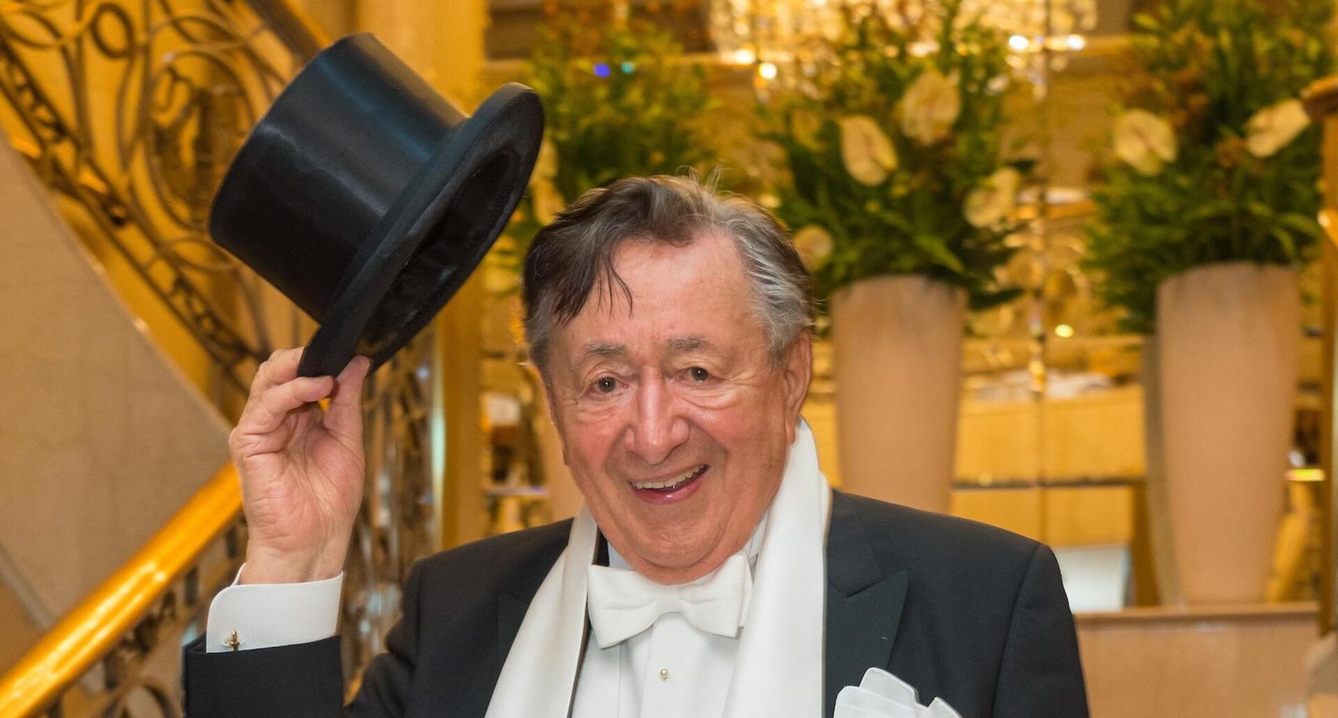 Richard Lugner in der wiener Staatsoper beim Opernball. Er trägt einen schwarzen Anzug und hält einen Hut in die Höhe.