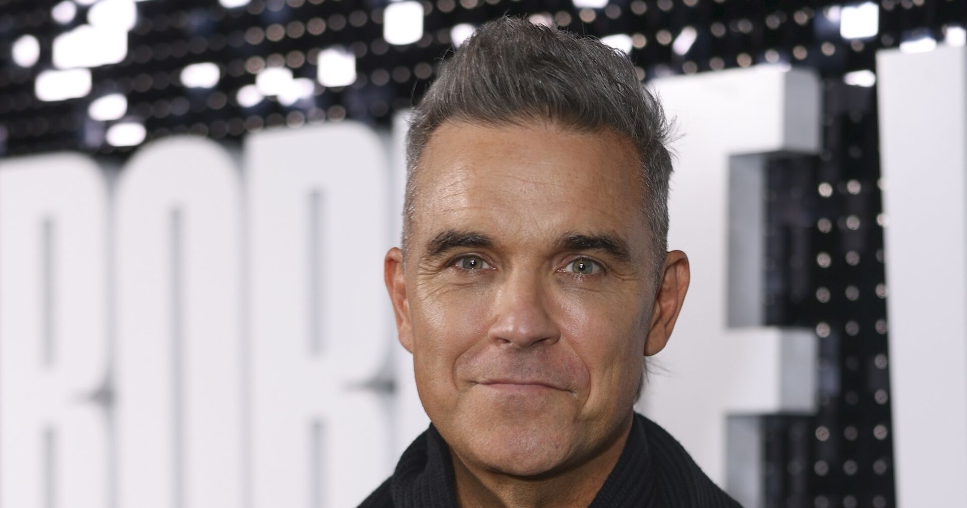 Robbie Williams mit aufgestellten grauen Haaren vor einer Fotoleinwand.