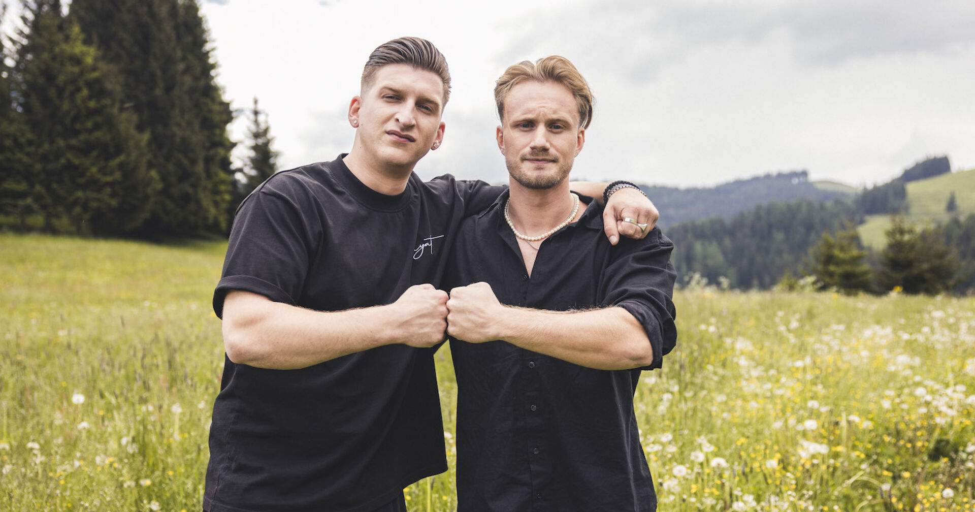 Satans Bratan und Mio tragen beide schwarze T-Shirts. Sie stehen vor einer Blumenwiese und geben sich siegessicher die Fäuste.