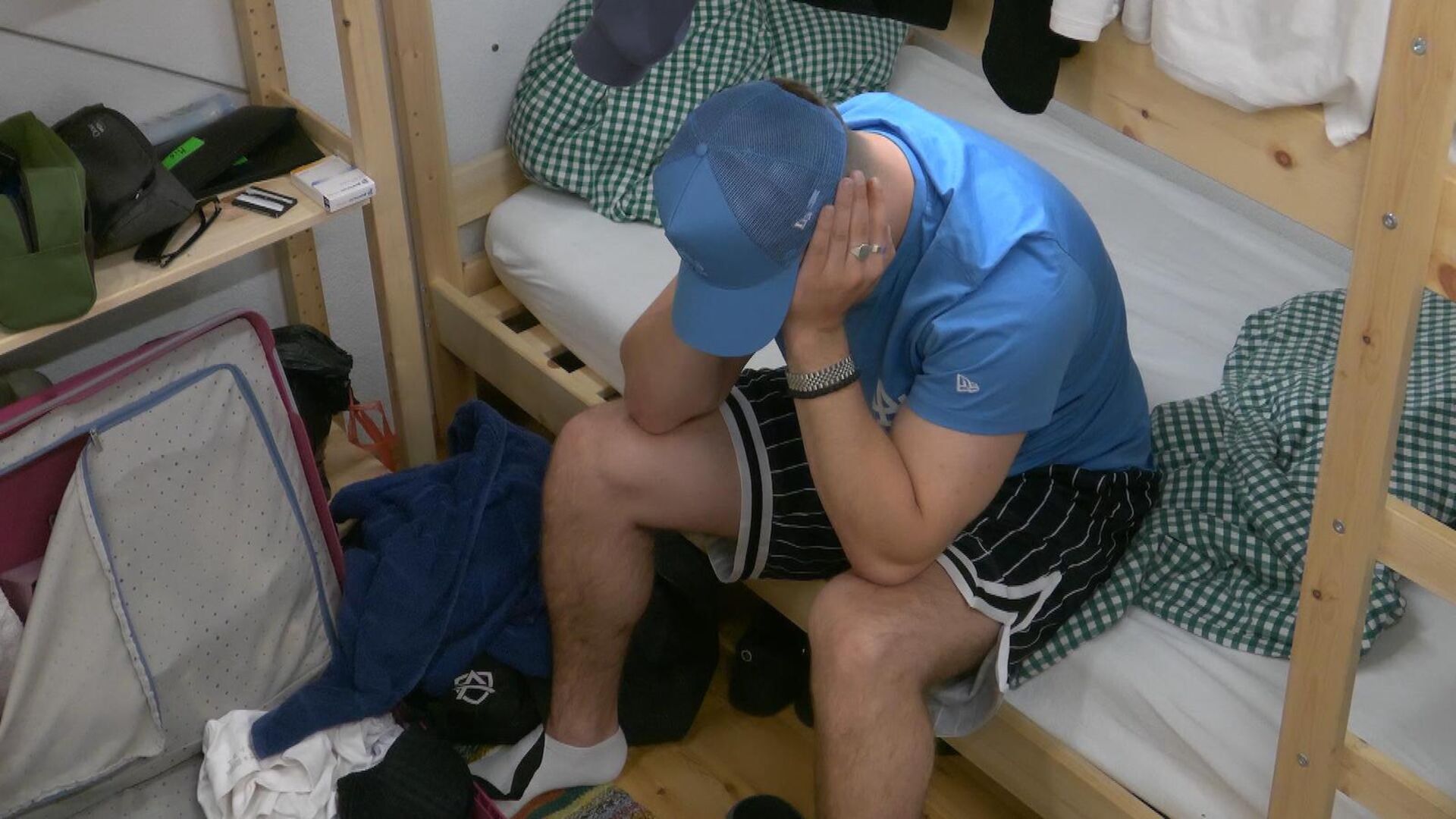 Satans Bratan sitzt mit einem blauen T-Shirt und einer blauen Kappe auf dem Bett und stützt sich nachdenklich in die Hände.