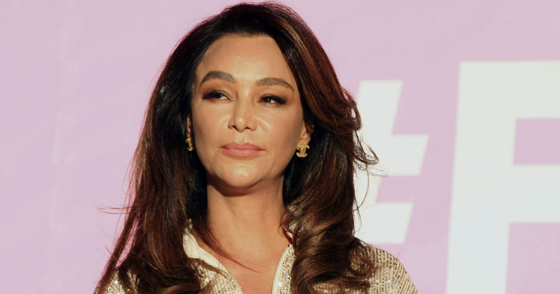 Verona Pooth trägt offene dunkle Haare und sitzt vor einer pinken Leinwand.