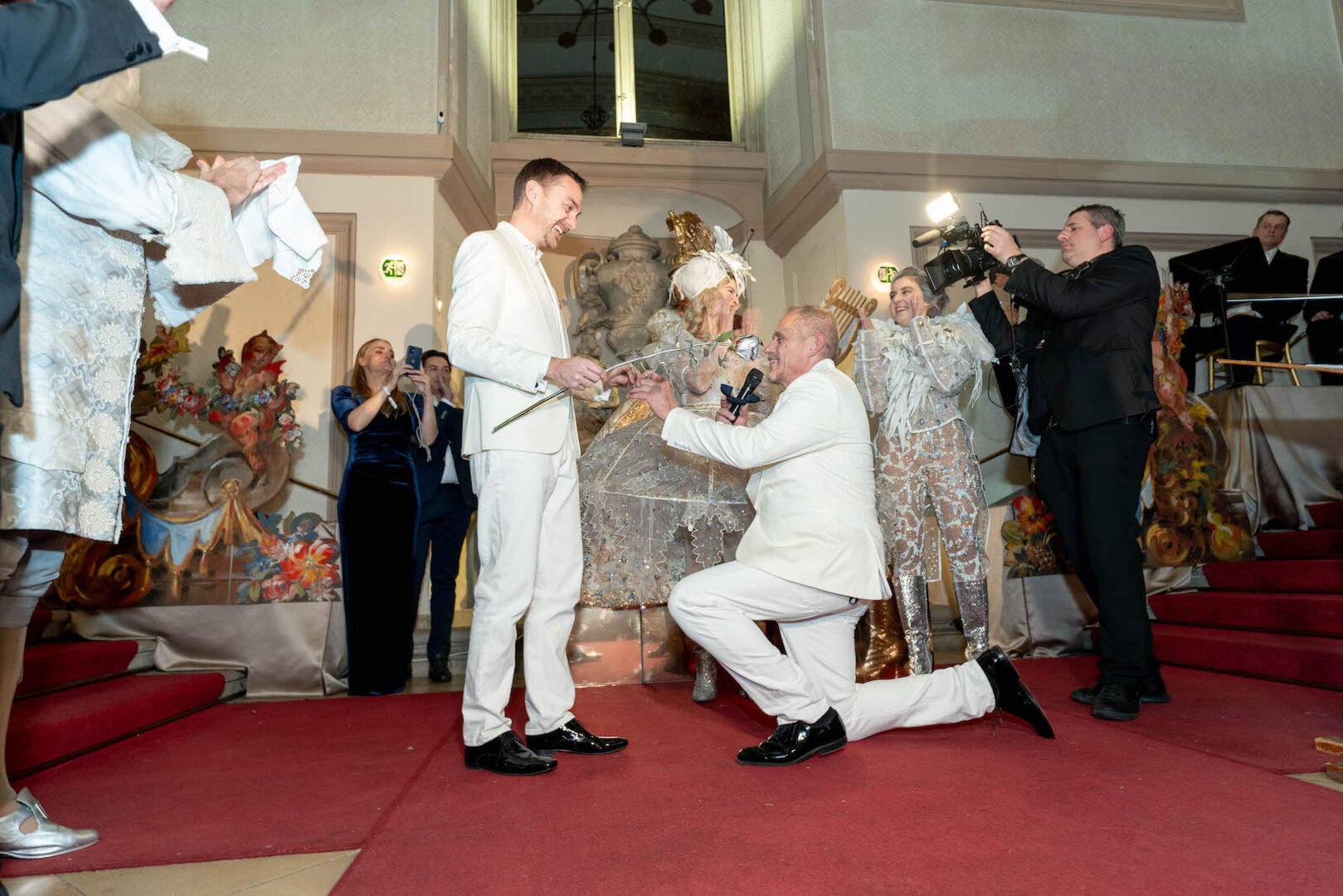 Gery Keszler und sein Verlobter tragen weiße Anzuge. Er ist auf den Knie und macht seinem Verlobten einen Heiratsantrag