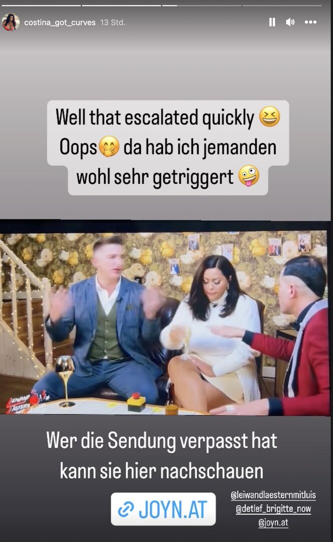 Satans Bratan und Costina sitzen auf einer Couch und gestikulieren heftig. Er trägt ein blaues Sakko und sie ein weißes Kleid.