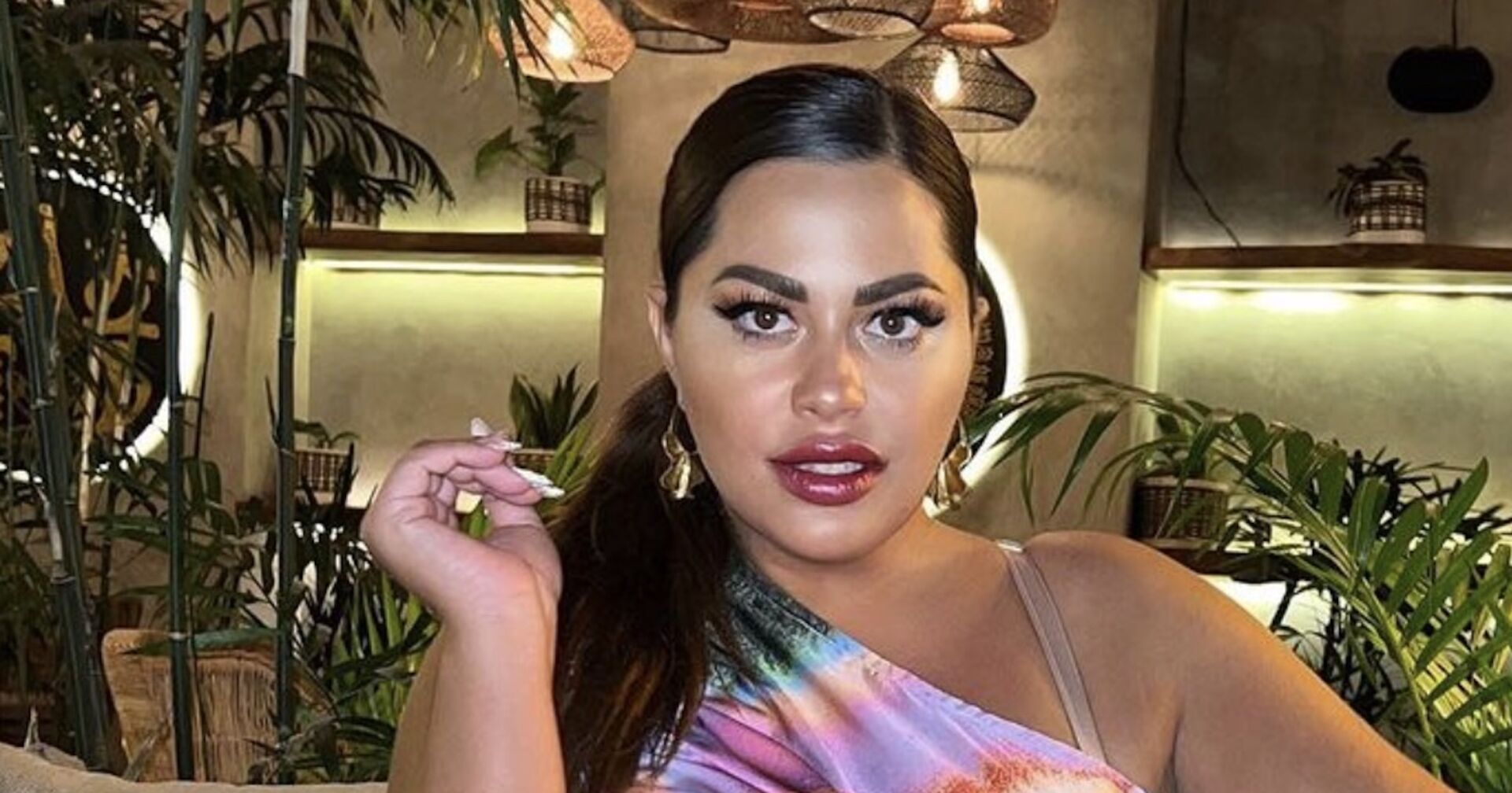 Costina mit einem Zopf und einem bunten Kleid. Im Hintergrund sind Pflanzen.