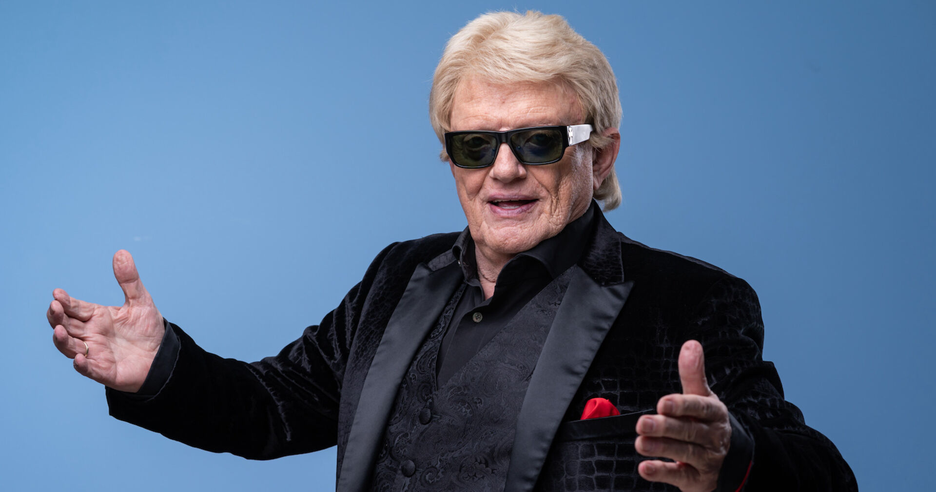 Heino steht vor einer blauen Fotowand mit einem schwarzen Sakko, blonden Haaren und einer Sonnenbrille.