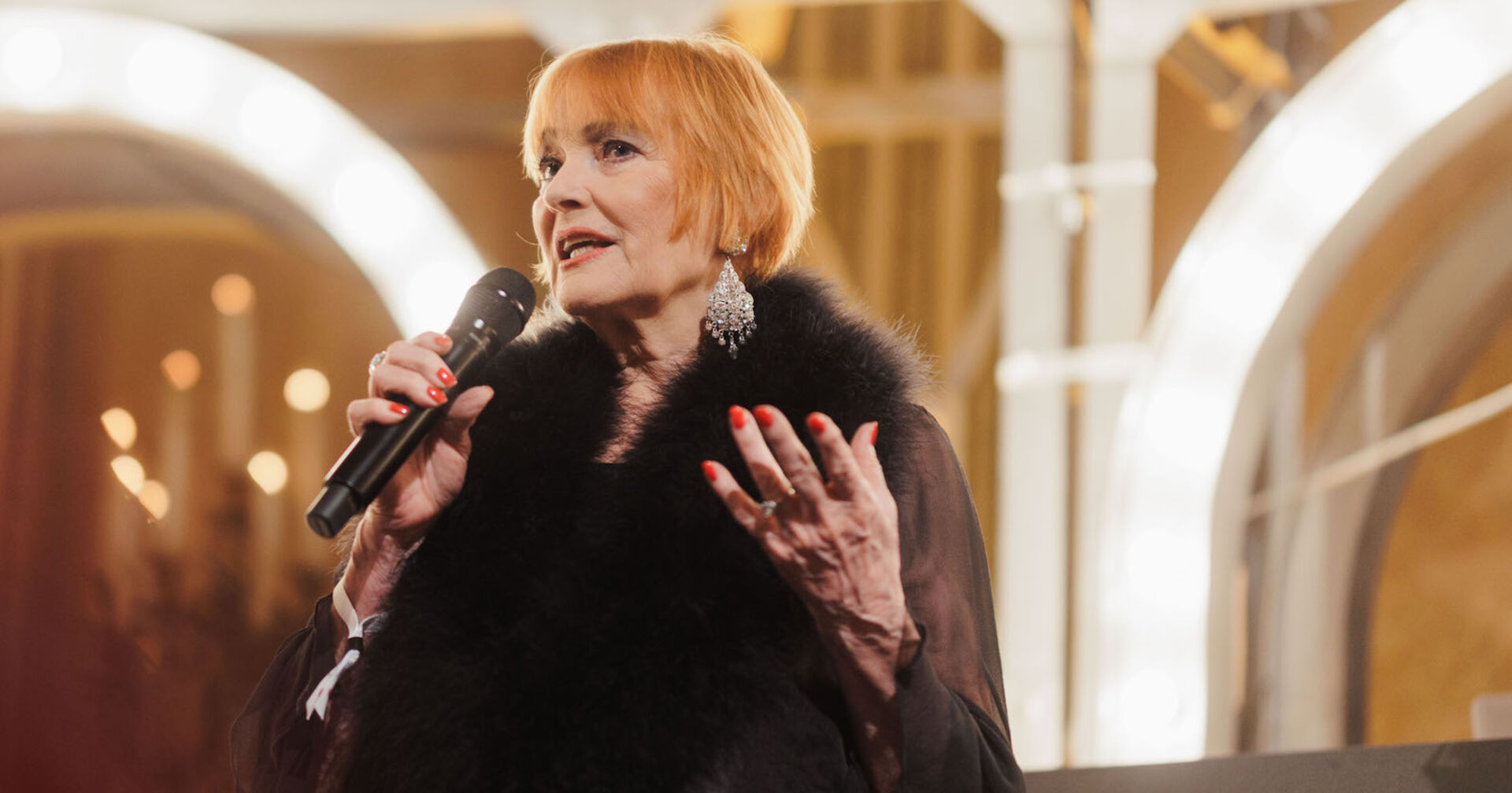 Chris Lohner mit kurzen roten Haaren und einem schwarzen Pelz. Sie spricht in ein Mikrofon.