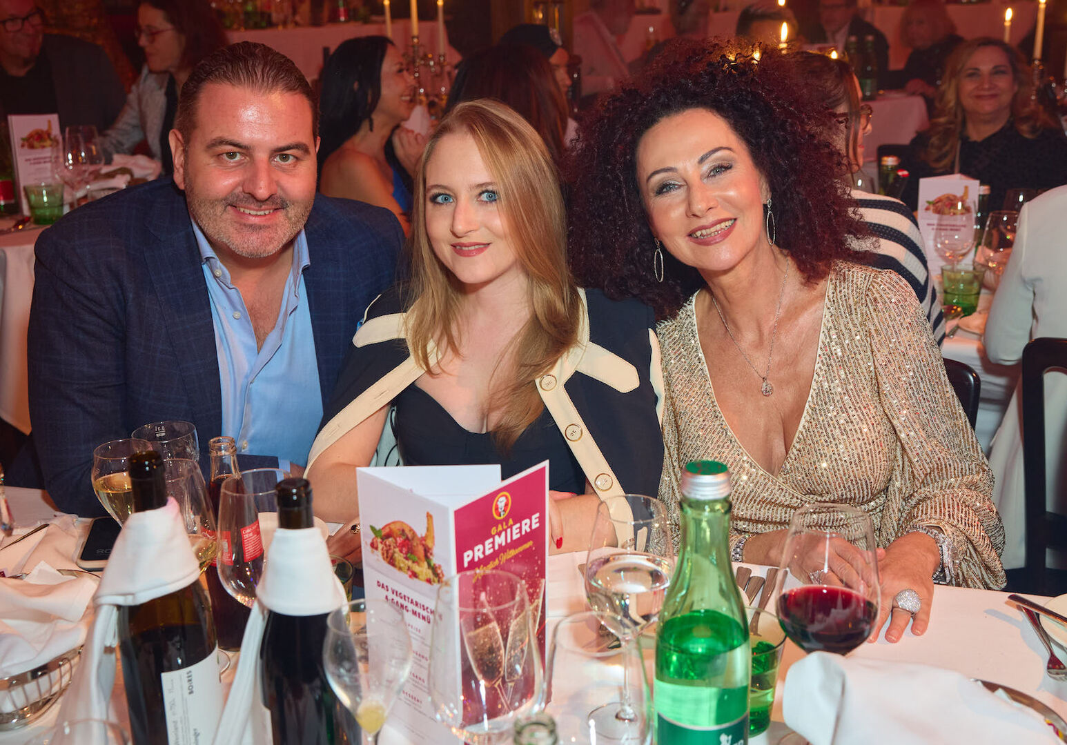 Leo Kohlbauer trägt ein hellblaues Hemd und ein schwarzes Sakko, Jaqueline Lugner ein dunkles Kleid und eine dunkle Jacke und Mausi Lugner ein gold-glitzer Kleid.