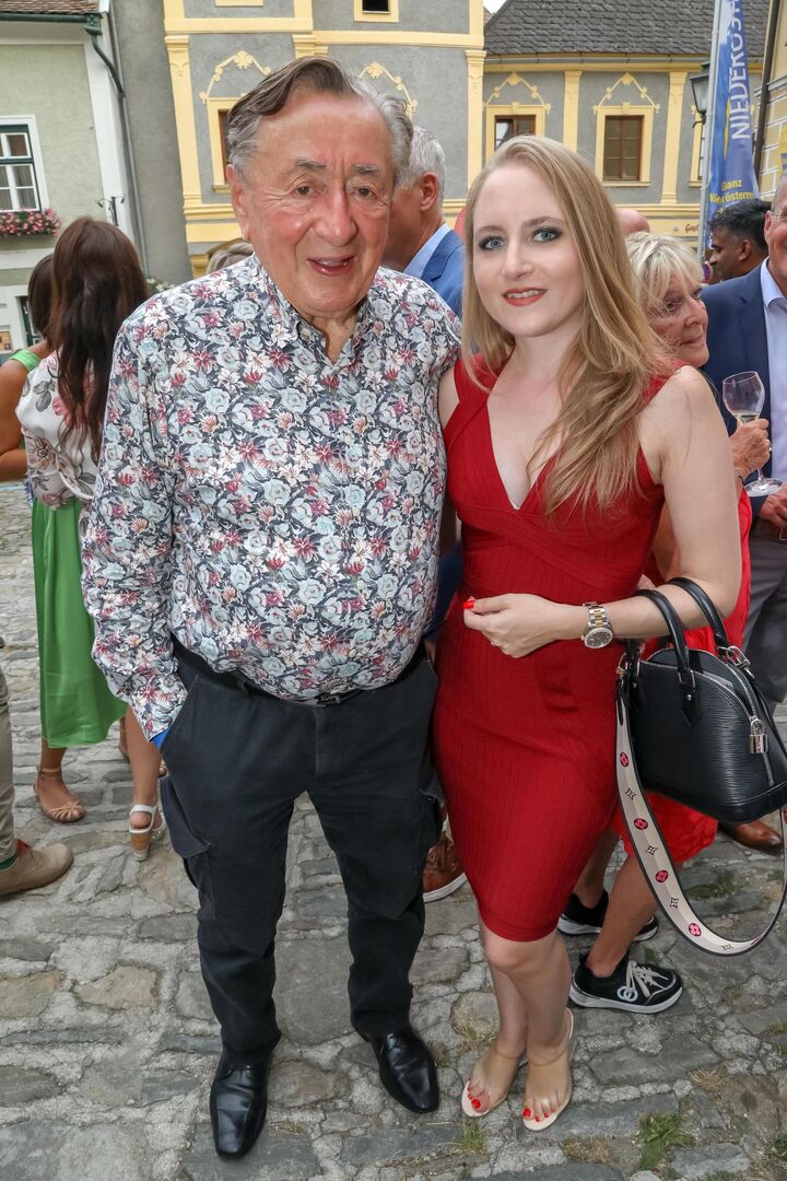 Richard Lugner trägt ein buntes Hemd und Jacky Lugner ein rotes Kleid und eine schwarze Tasche.