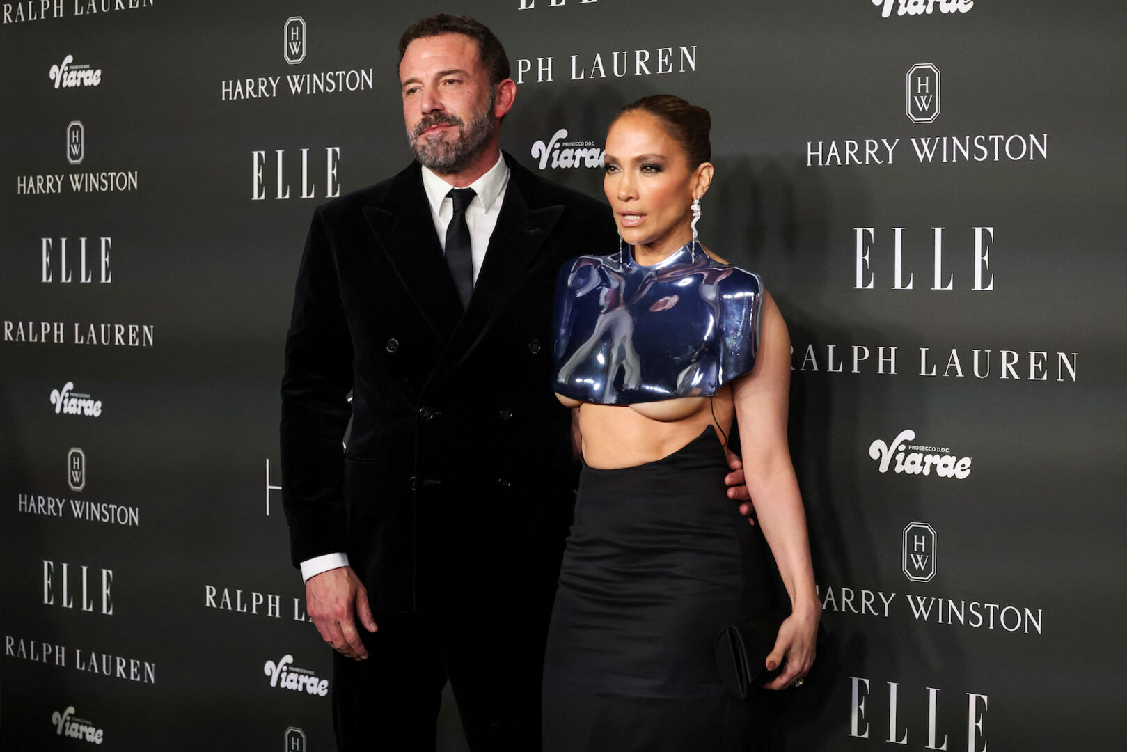 Jennifer Lopez trägt ein metallenes Oberteil und einen Bleistiftrock. Ben Affleck trägt einen schwarzen Anzug.