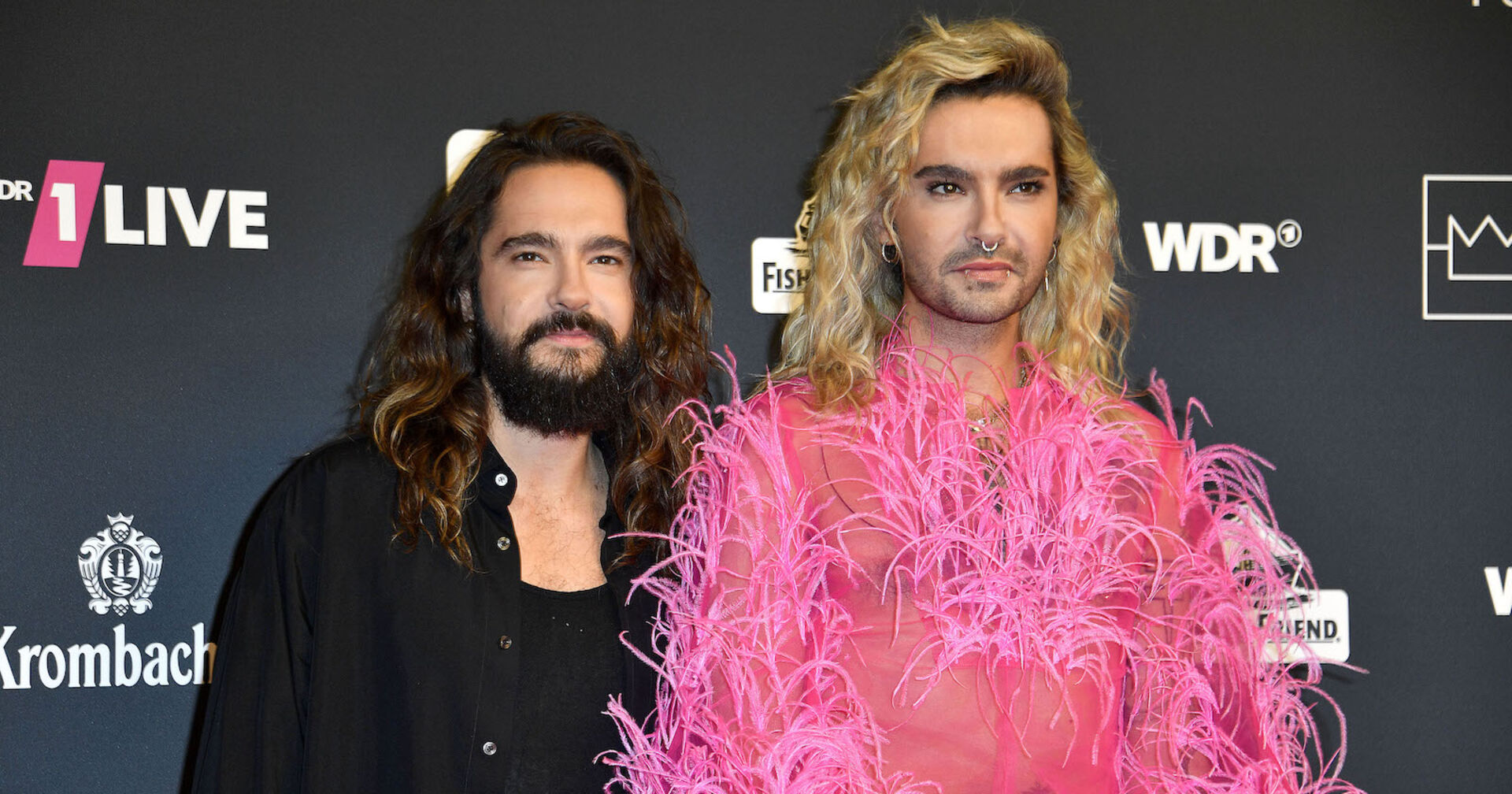 Die Kaulitz Twins. Tom trägt ein schwarzes Hemd und Bill einen rosa Federn Anzug.