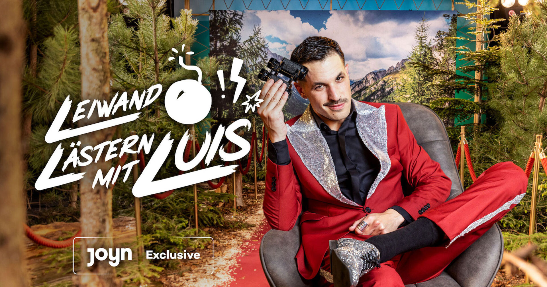 Luis mit einem roten Anzug und einem schwarzen Hemd. Er hält ein Fernglas in der Hand.