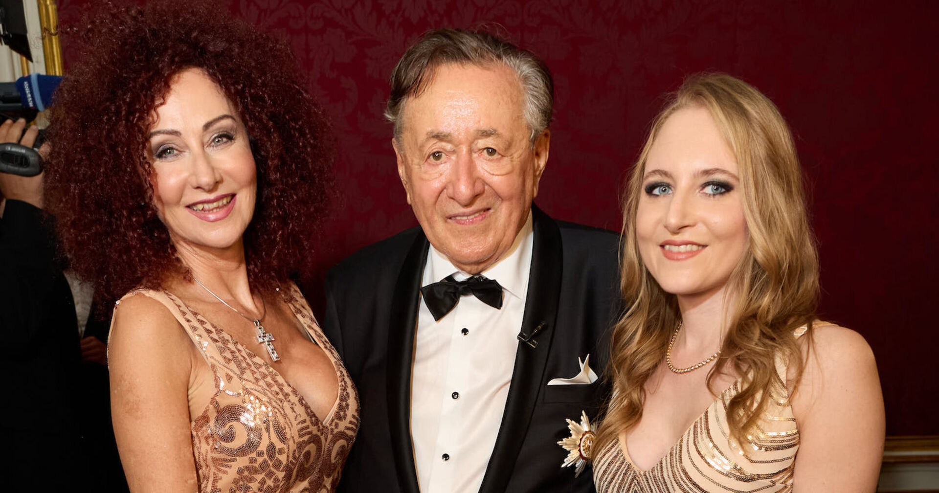 Christine Lugner trägt ein goldenes Kleid und offene Haare, Richard Lugner einen Smoking und Jacky Lugner ein Ballkleid und offene blonde Haare.