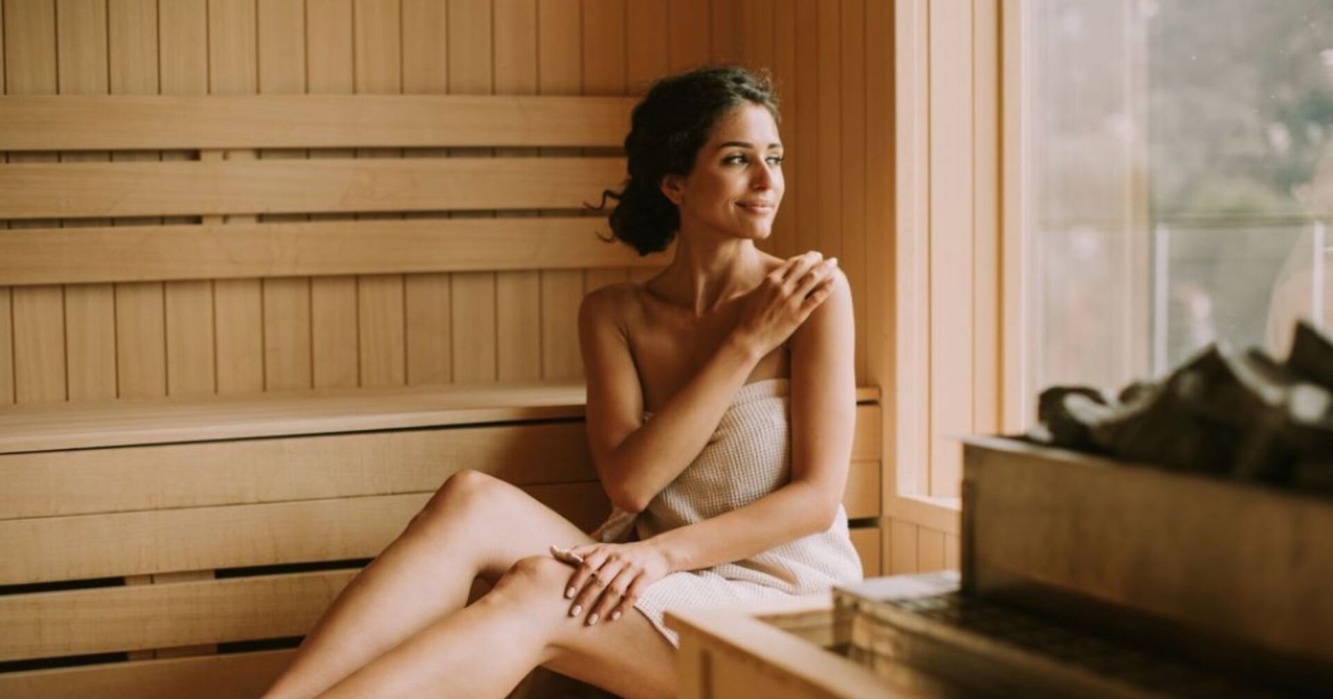 Eine Frau sitzt mit einem Handtuch bedeckt in einer gemütlichen Sauna aus Holz und schaut glücklich aus dem Fenster neben ihr.