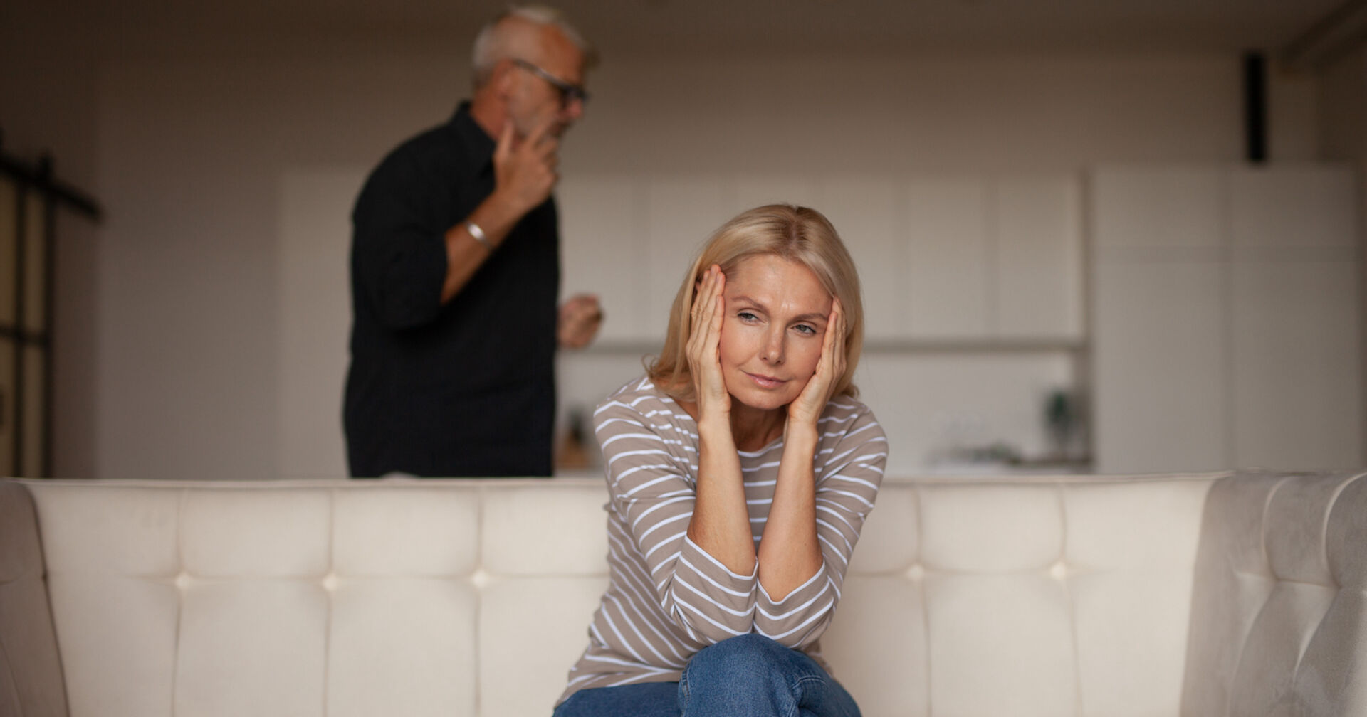 Ein älterer Mann streitet mit seiner Frau. Sie sitzt auf der Couch und hält sich die Ohren zu
