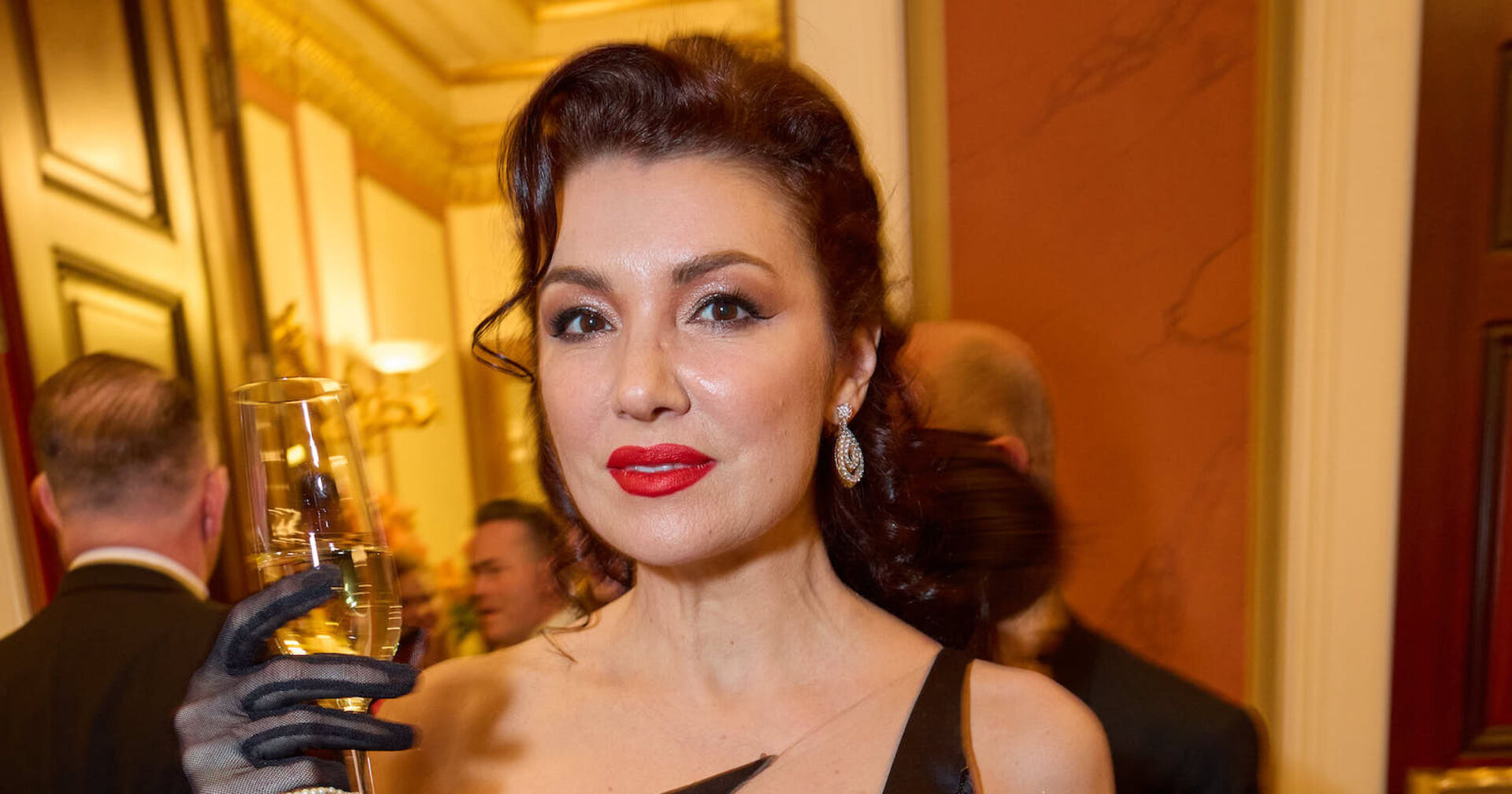 Anna Netrebko mit einer Ballrobe und einem Champagner-Glas in der Hand. Sie trägt roten Lippenstift.