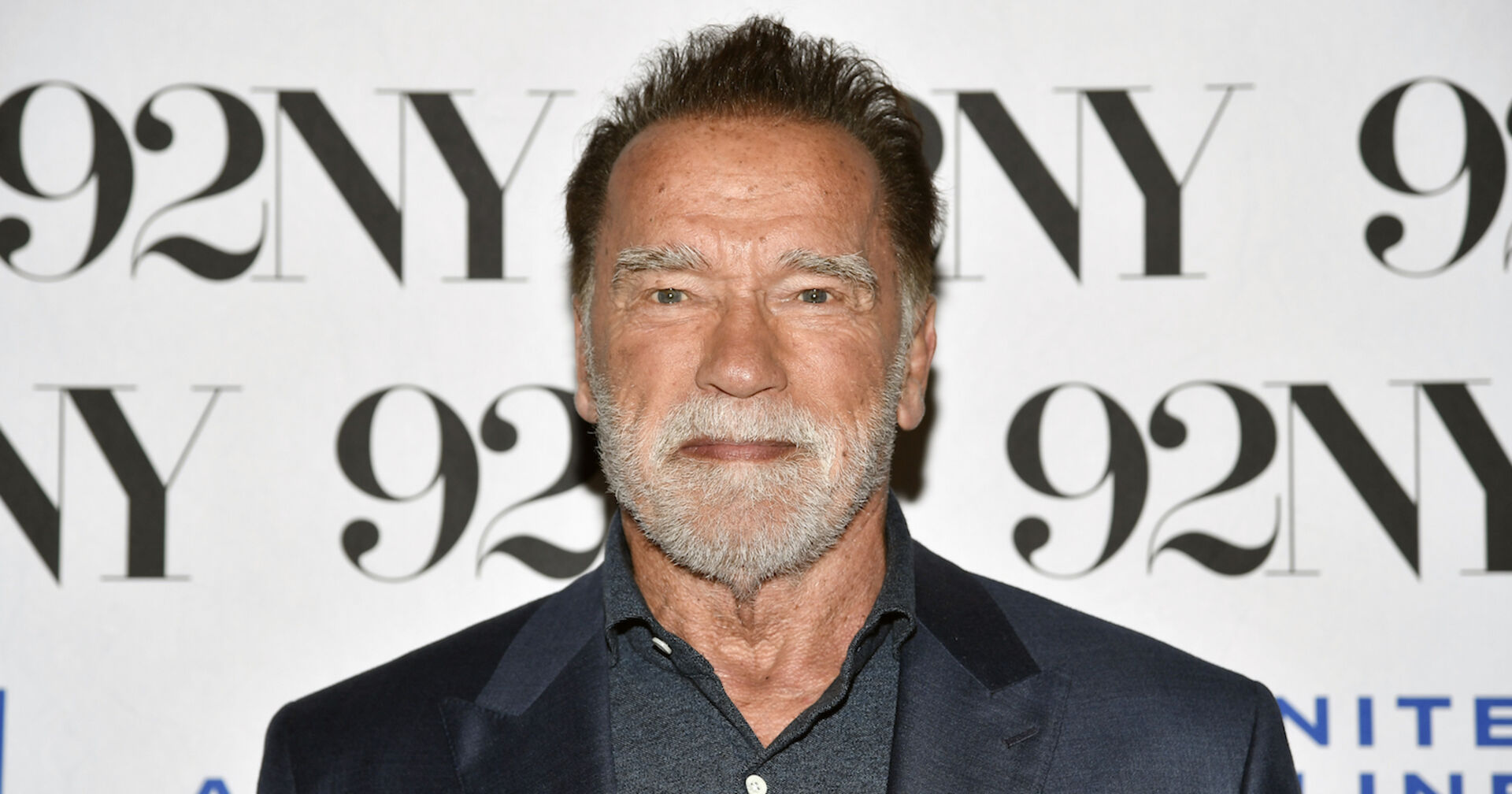 Arnold Schwarzenegger mit einem dunkelgrauen Smoking vor einer Fotowand.