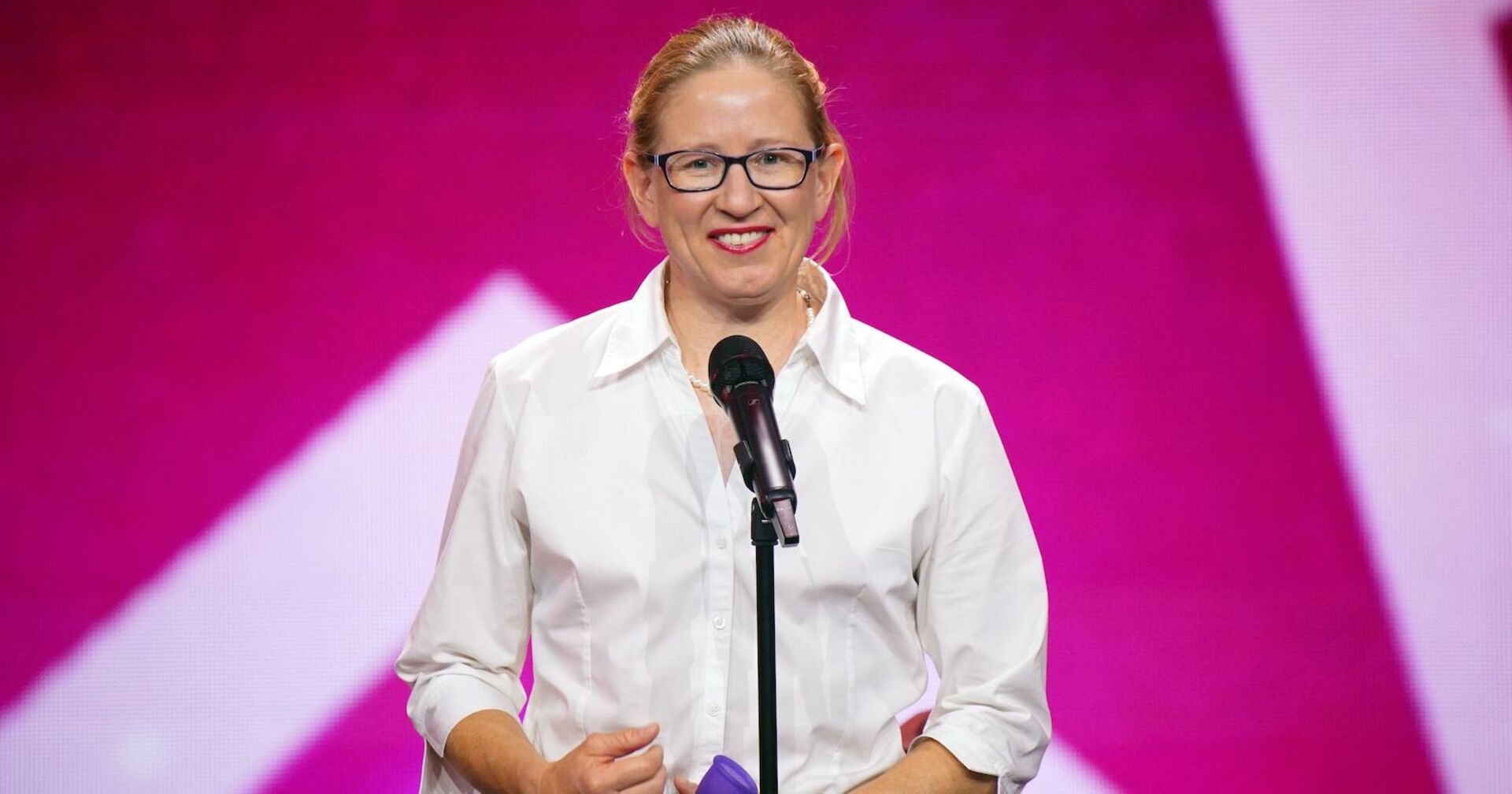 Die australische &quot;Das Supertalent&quot; Teilnehmerin Beatrice McQueef steht vor einem Mikrofon und trägt ein weißes Hemd. Hinter ihr ist eine pinke Fotowand.