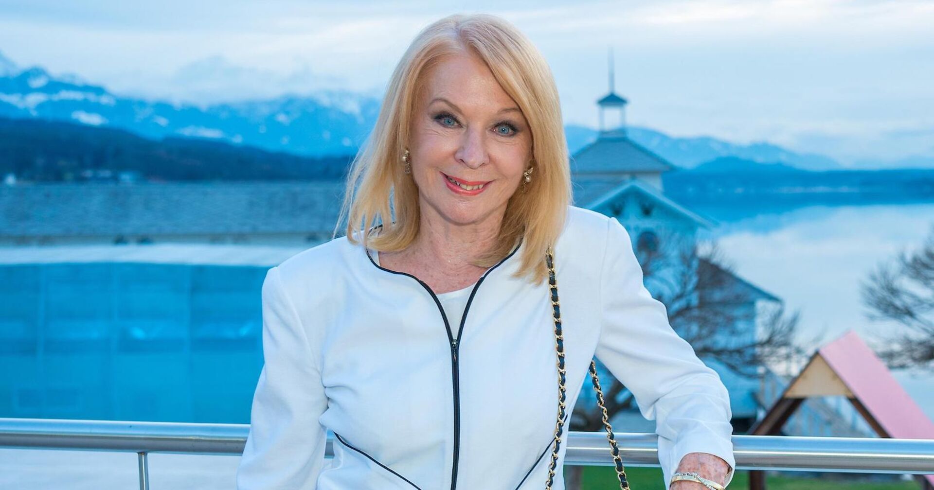 Gerda Rogers auf einem Balkon. Hinter ihr ist ein See und Berge. Sie trägt eine weiße Jacke und offene blonde Haare.