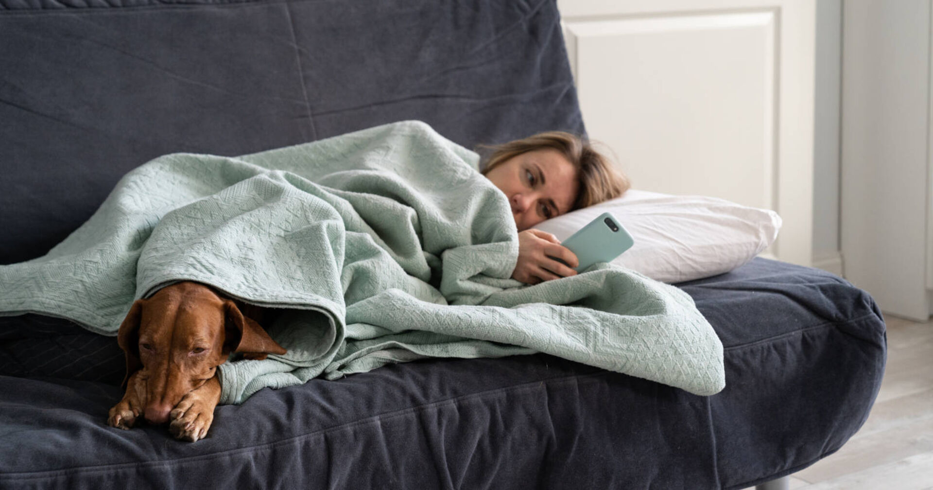 Eine Frau liegt motivationslos mit ihrem Hund unter der Decke.