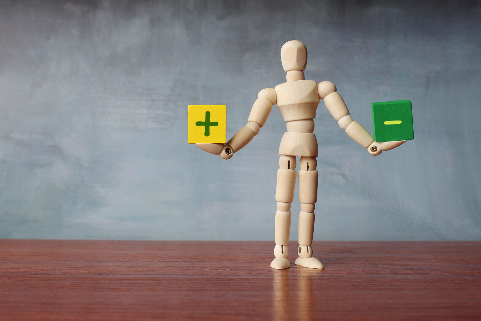 Eine Holzpuppe, die einen gelben Würfel mit einem grünen Plus sowie einen grünen Würfel mit einem weißen Minus in Händen hält