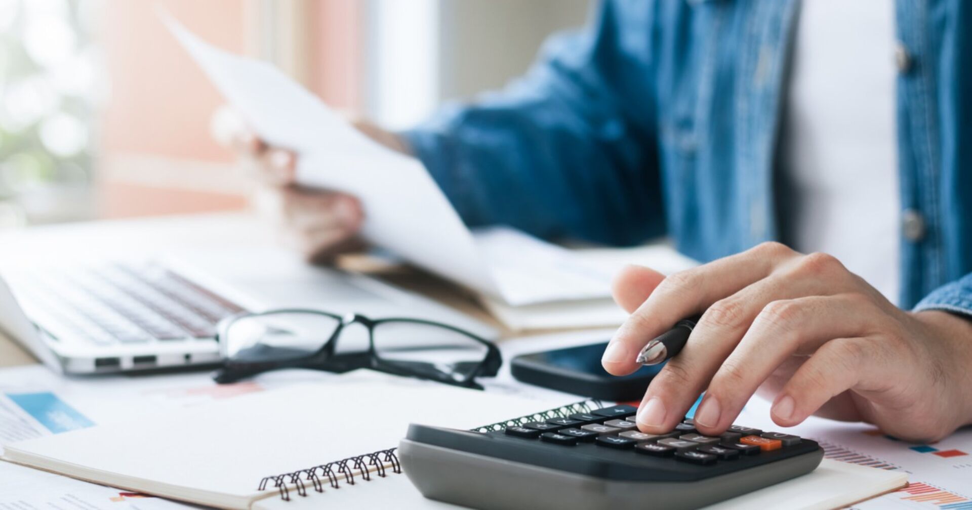 Mann sitzt vor seinem Laptop und hält einen Zettel in seiner Hand während er auf einem Taschenrechner tippt.