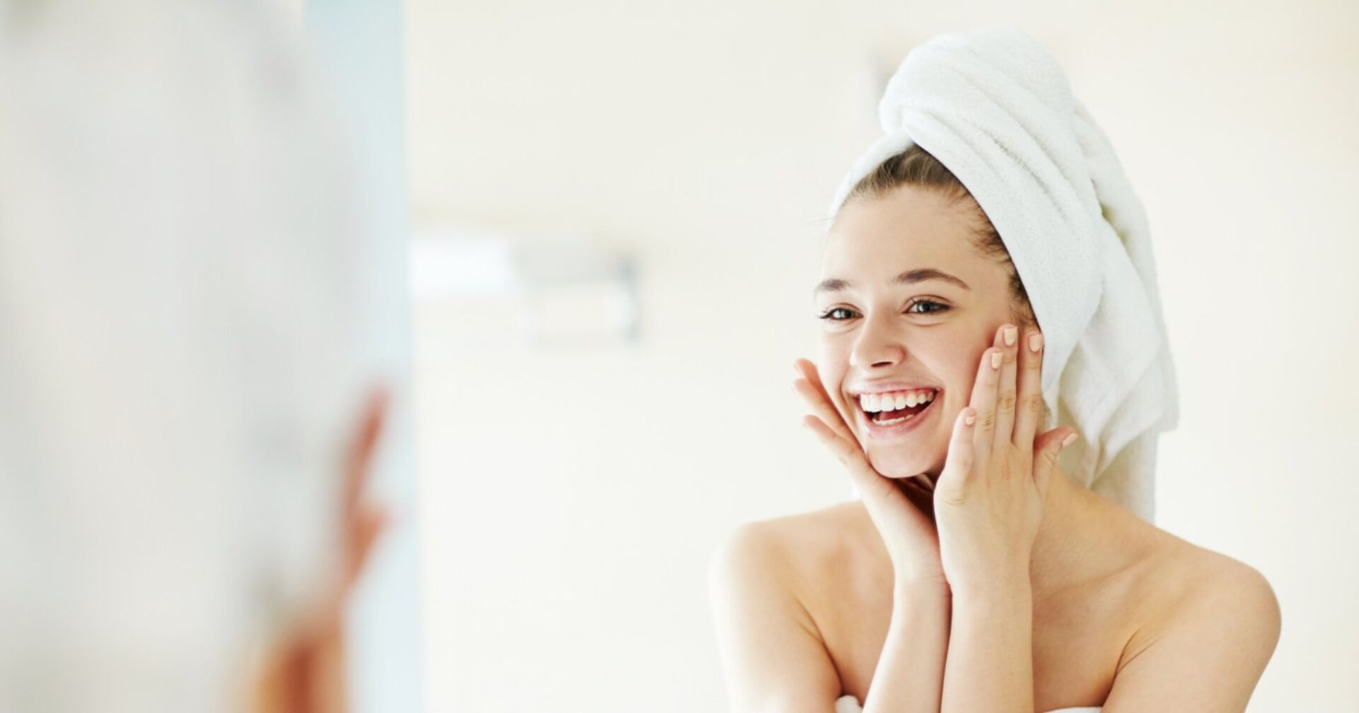 Eine junge Frau steht mit ihren Haaren in ein Handtuch eingewickelt vor dem Spiegel, lächelt und hält ihre Hände an ihre Wangen