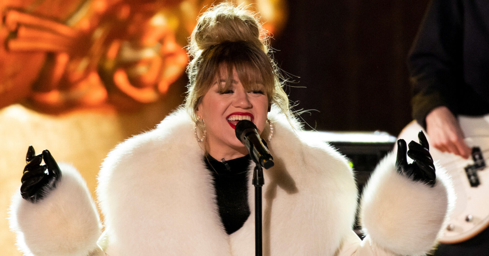 Kelly Clarkson trägt einen weißen Pelzmantel und steht auf einer Bühne.