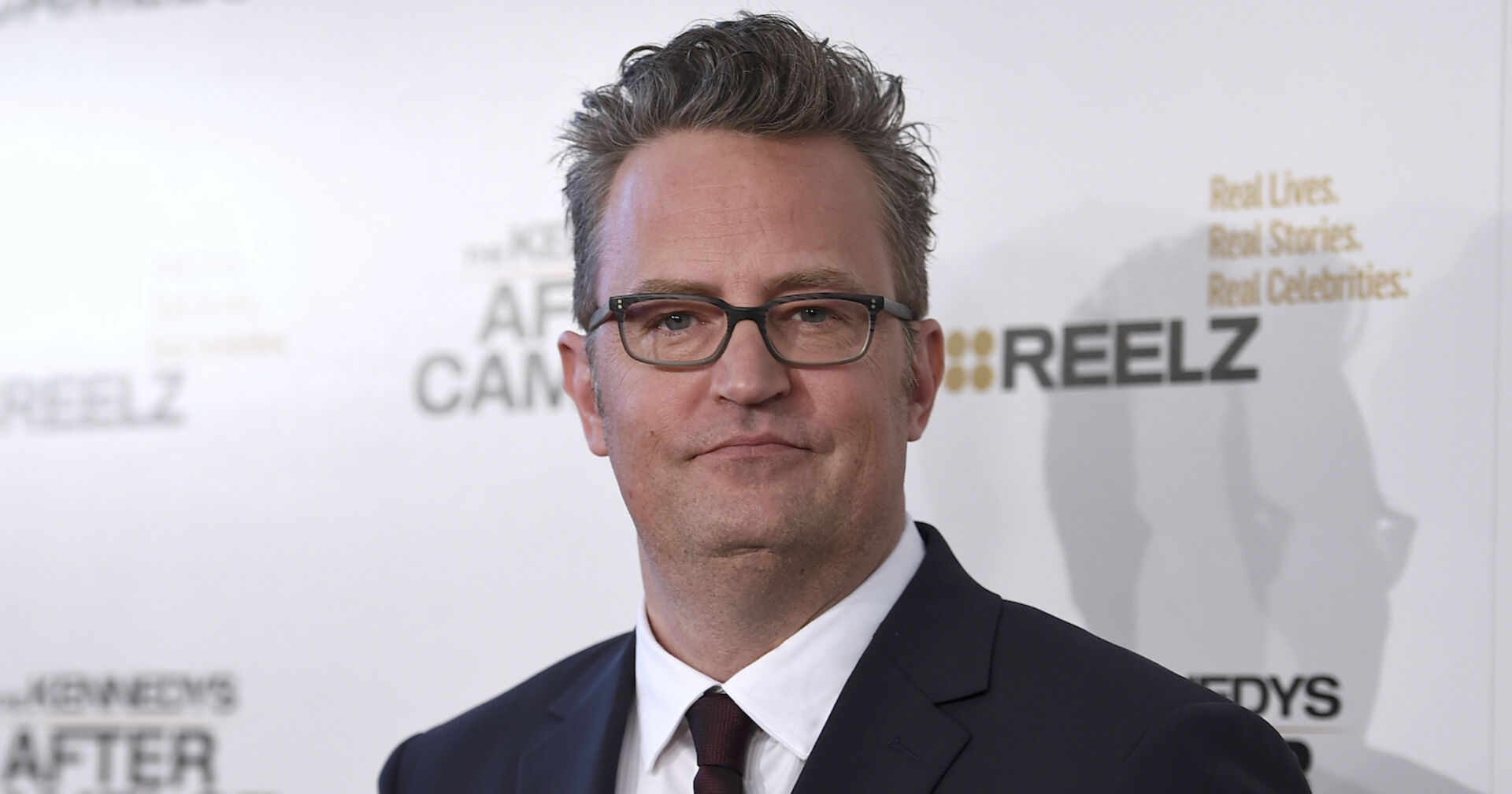 Matthew Perry vor einer Fotoleinwand mit einem Sakko.