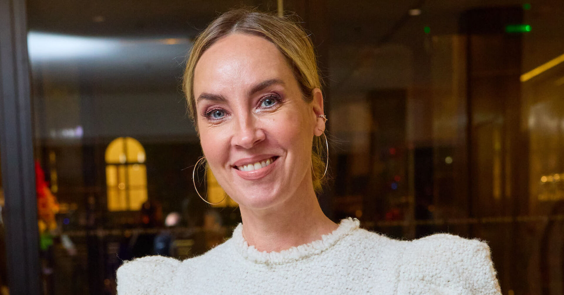 Nadja Bernhard in einem weißen Pullover mit geschlossenen Haaren und großen Ohrringen.