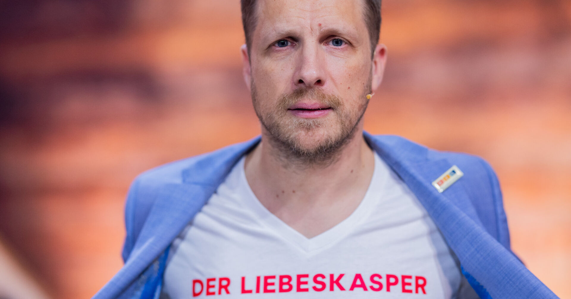 Oliver Pocher mit einem weißen T-Shirt auf dem steht &quot;Liebeskasper&quot; und einem hellblauen Sakko.