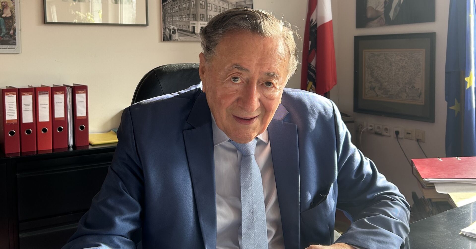 Richard Lugner auf seinem Schreibtisch. Er trägt einen blauen Anzug und eine Krawatte.