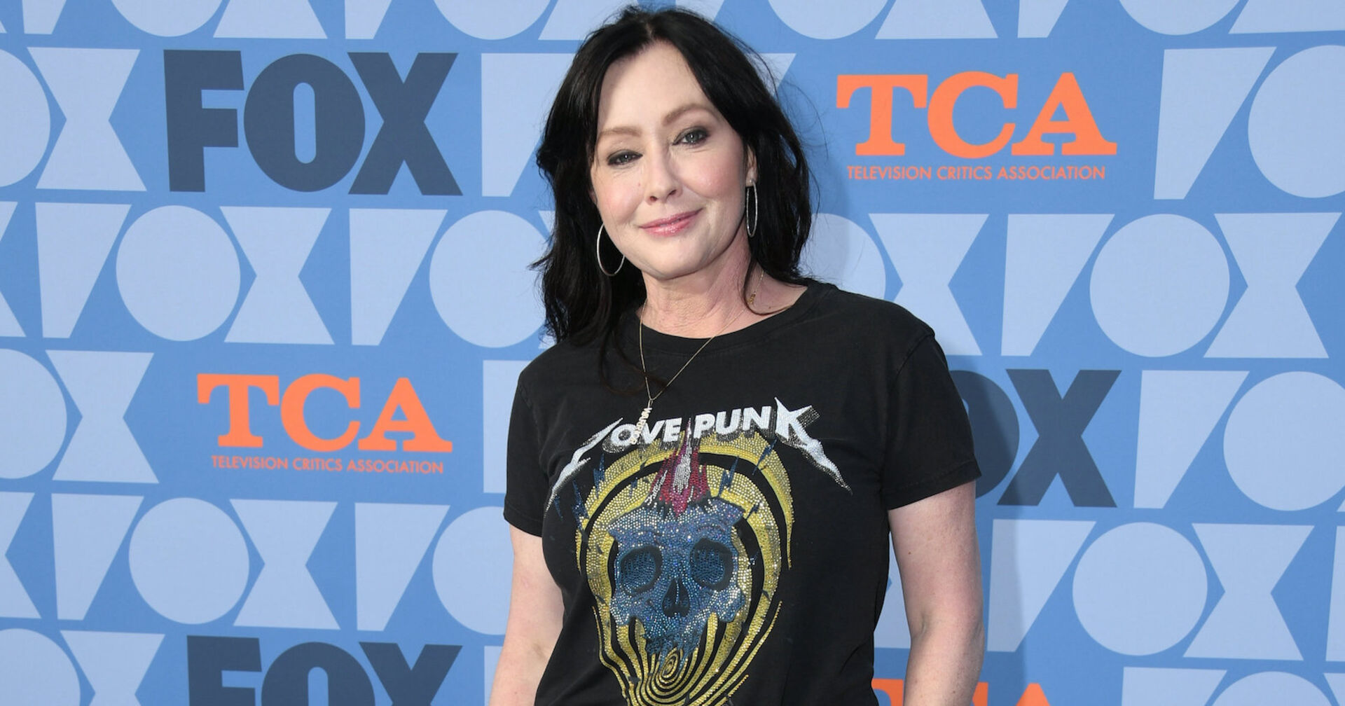 Shannen Doherty vor einer Fotoleinwand mit einem schwarzen T-Shirt.