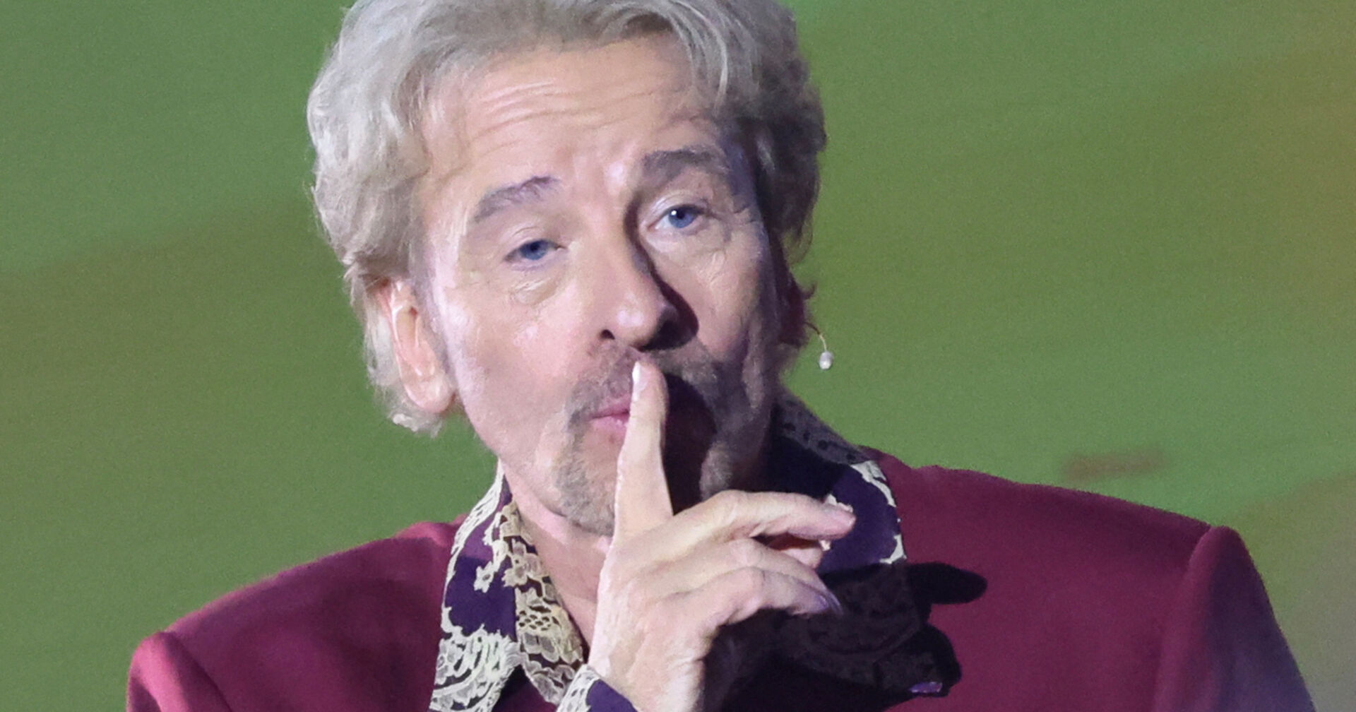Thomas Gottschalk hält sich den Zeigefinger schweigend vor den Mund. Er trägt ein weinrotes Sakko.