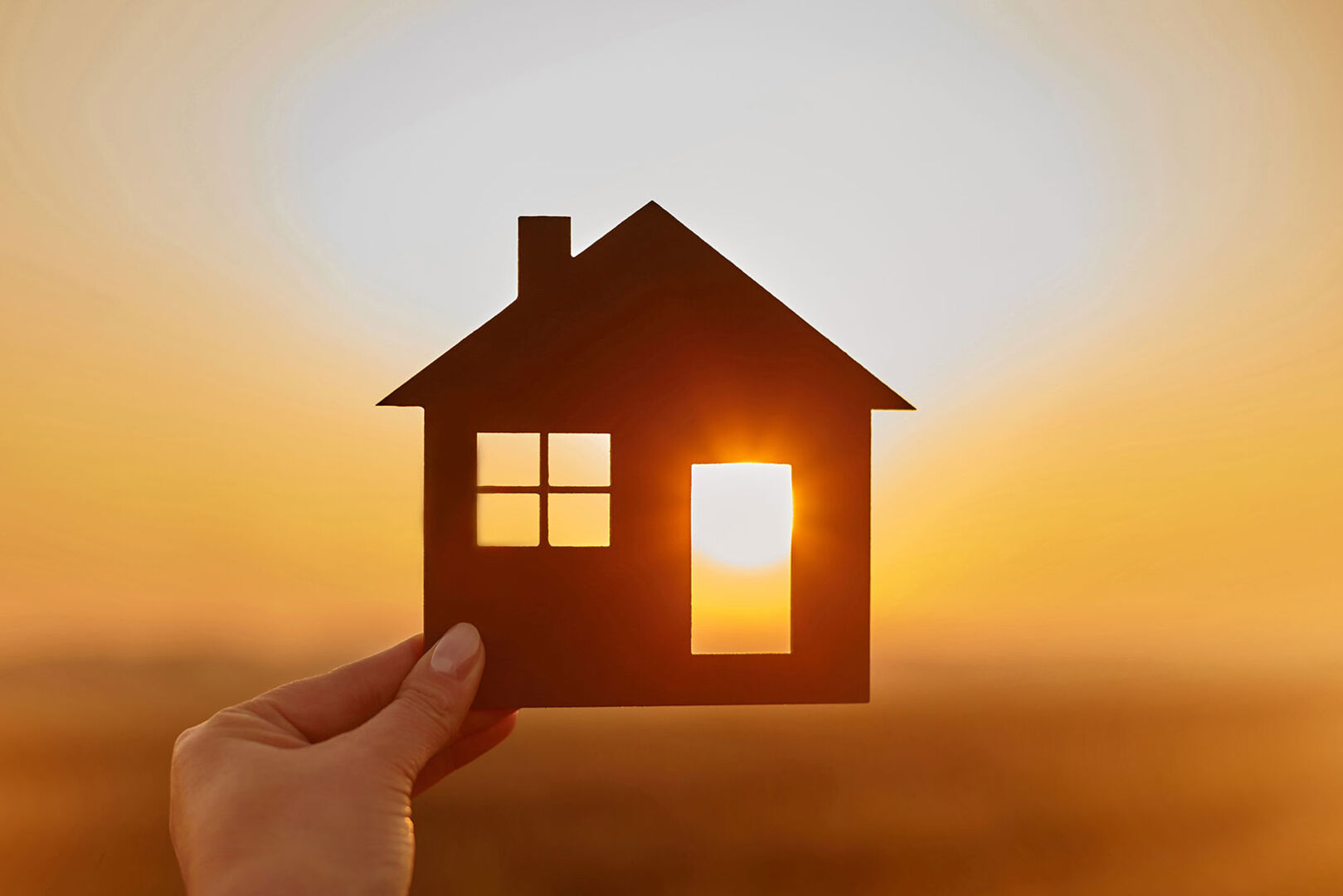 Junge Frau hält ein Miniaturhaus gegen das Licht der untergehenden Sonne | Credit: iStock.com/bodnarchuk
