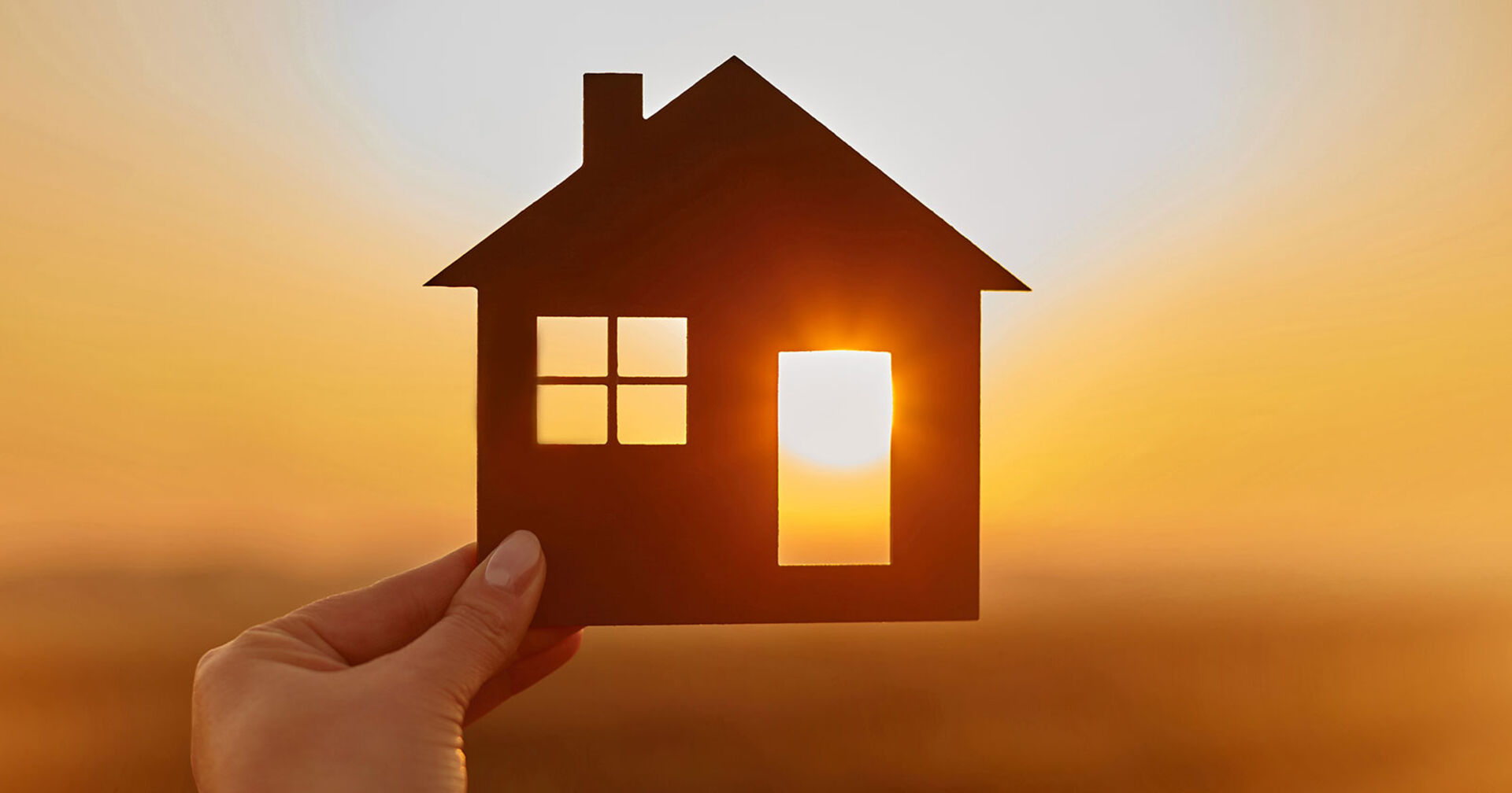 Junge Frau hält ein Miniaturhaus gegen das Licht der untergehenden Sonne | Credit: iStock.com/bodnarchuk