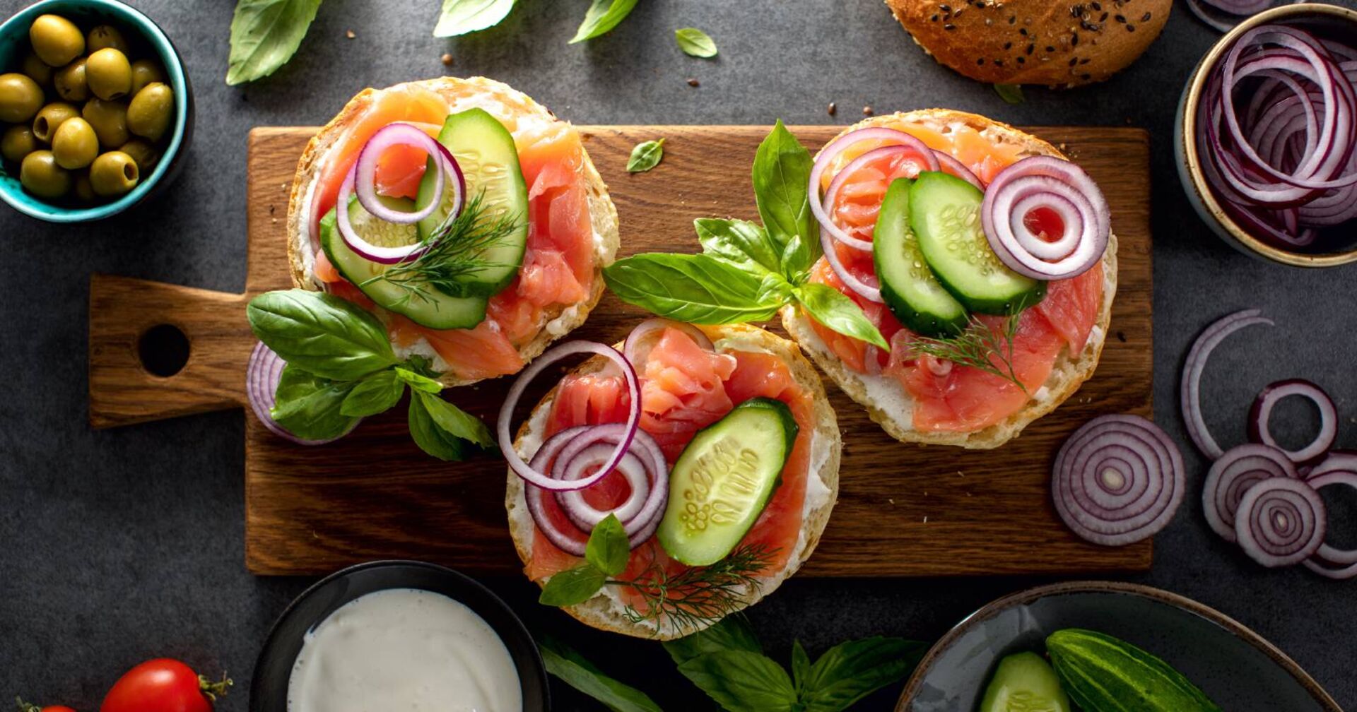 Auf einem Holzbrett liegen 3 Bagels, belegt mit Lachs, roten Zwiebelringen und Gurkenscheiben.