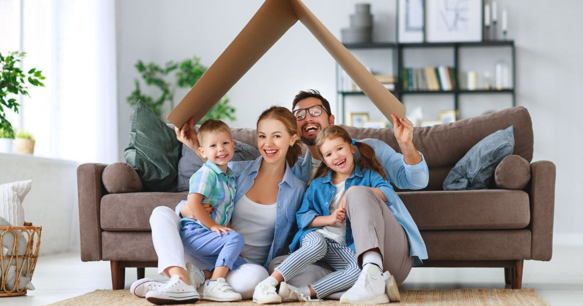 Vierköpfige Familie sitzt im Wohnzimmer am Boden und formt mit einem Karton ein Dach über ihren Köpfen.